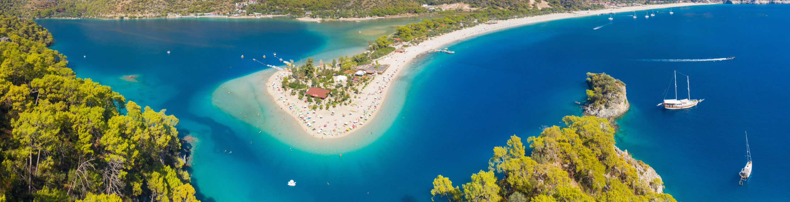 Stunning Blue Lagoon Beach in Turkey An aerial view of a unique sandy peninsula with white beaches extending into turquoise blue waters, surrounded by green forested coastline and featuring several boats and sailboats.