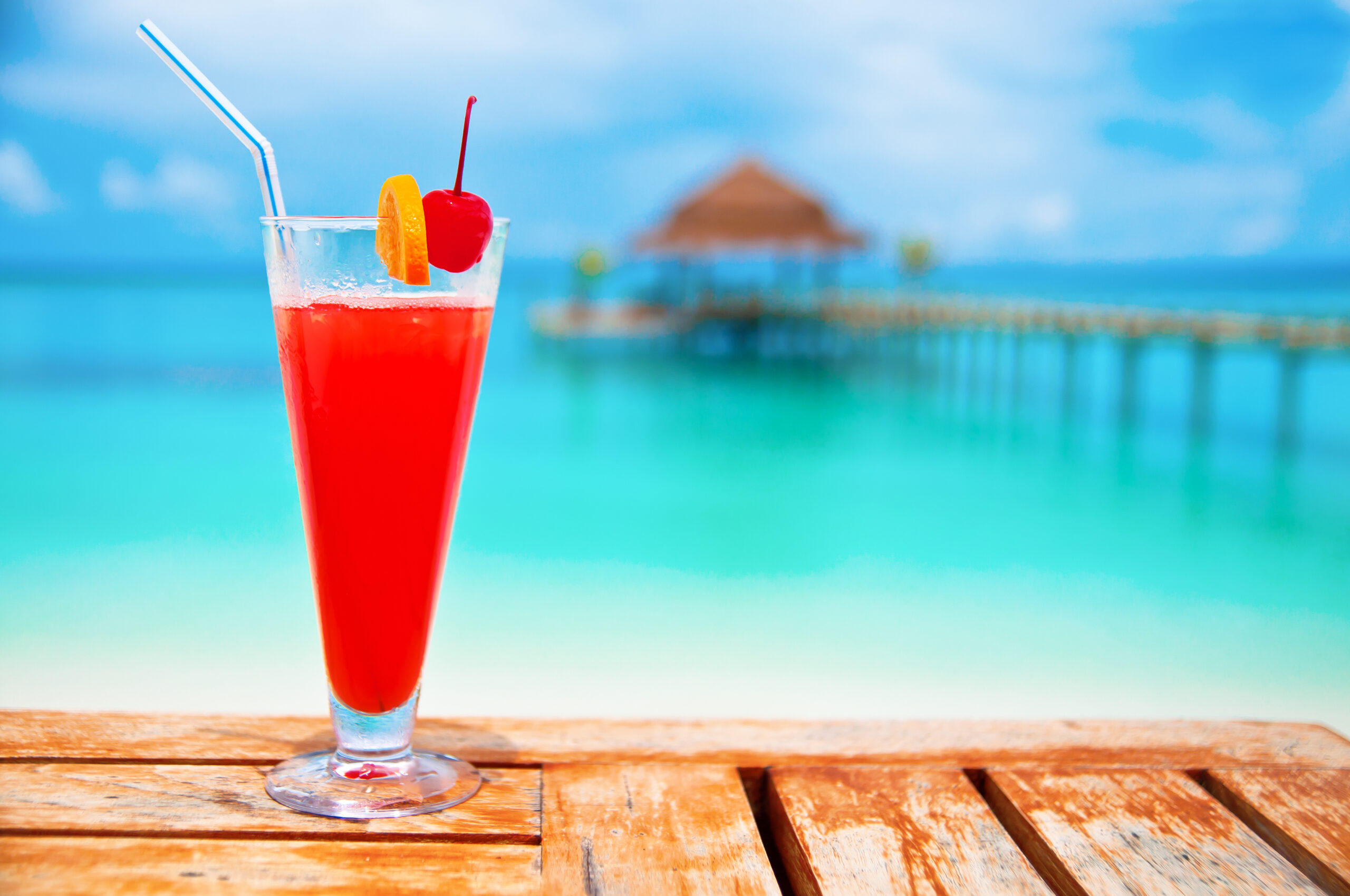 Tropical Beach Cocktail with Ocean View A vibrant red cocktail with ice, garnished with a maraschino cherry and orange slice, sits on a wooden deck overlooking turquoise tropical waters.
