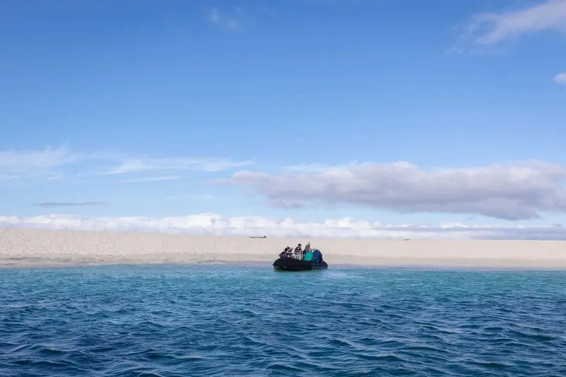 Zodiac Landing Craft in Arctic Waters A small Zodiac landing craft with passengers approaches across turquoise Arctic waters, with a snow-covered mountainous coastline visible in the background under a partly cloudy sky.