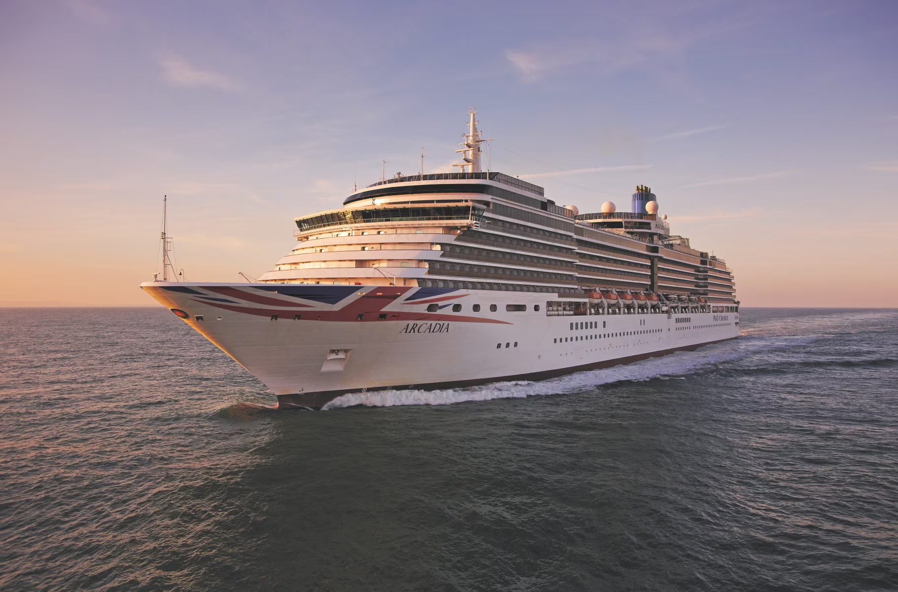 The P&O Arcadia, a large modern cruise ship, is pictured in shallow coastal waters at golden hour. The vessel displays the distinctive P&O livery with Union Jack design on its hull, with multiple decks visible against a serene twilight sky and calm sea.