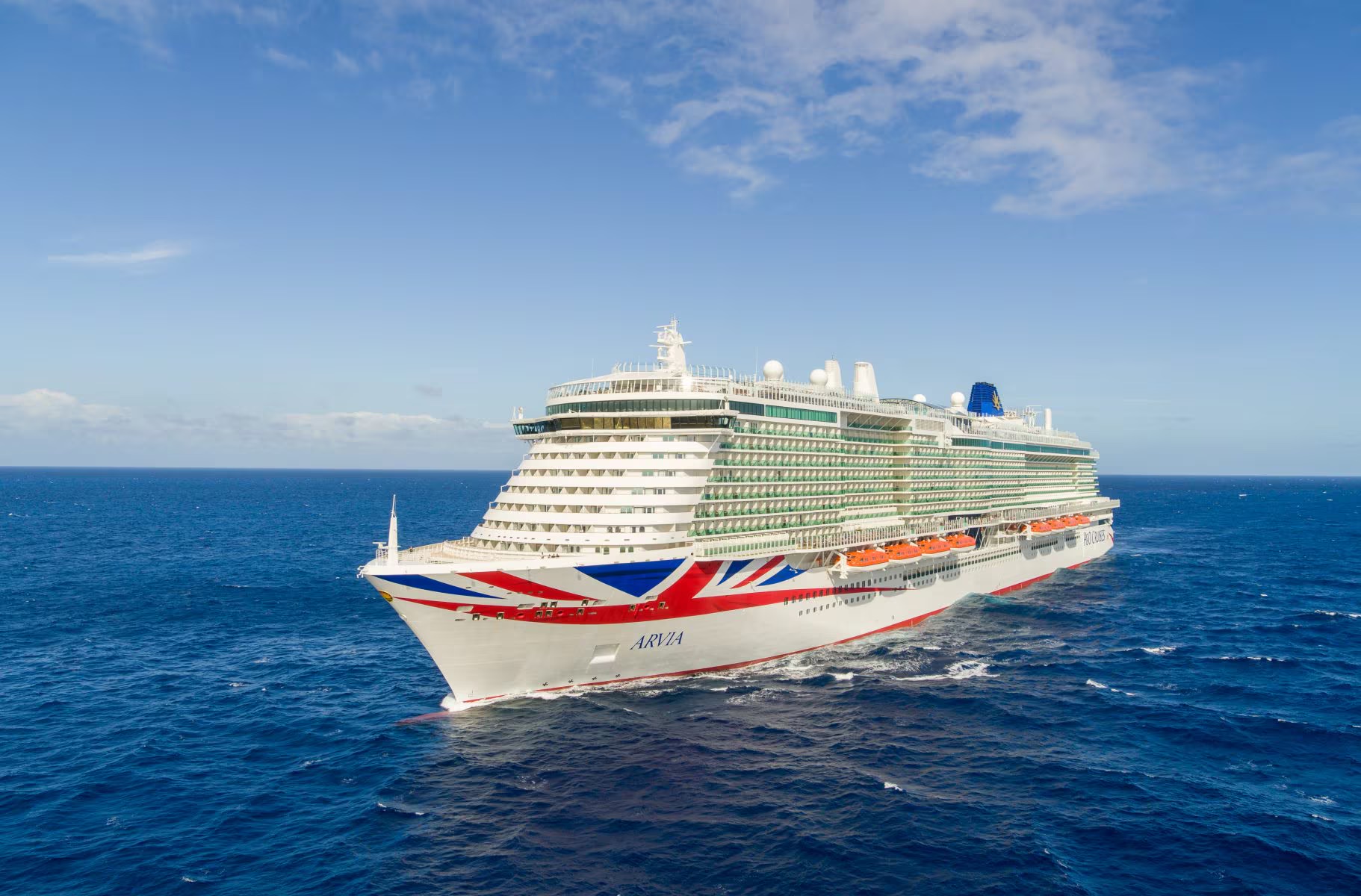A modern large cruise ship with distinctive white hull and Union Jack-inspired livery sails through deep blue ocean waters under clear skies.