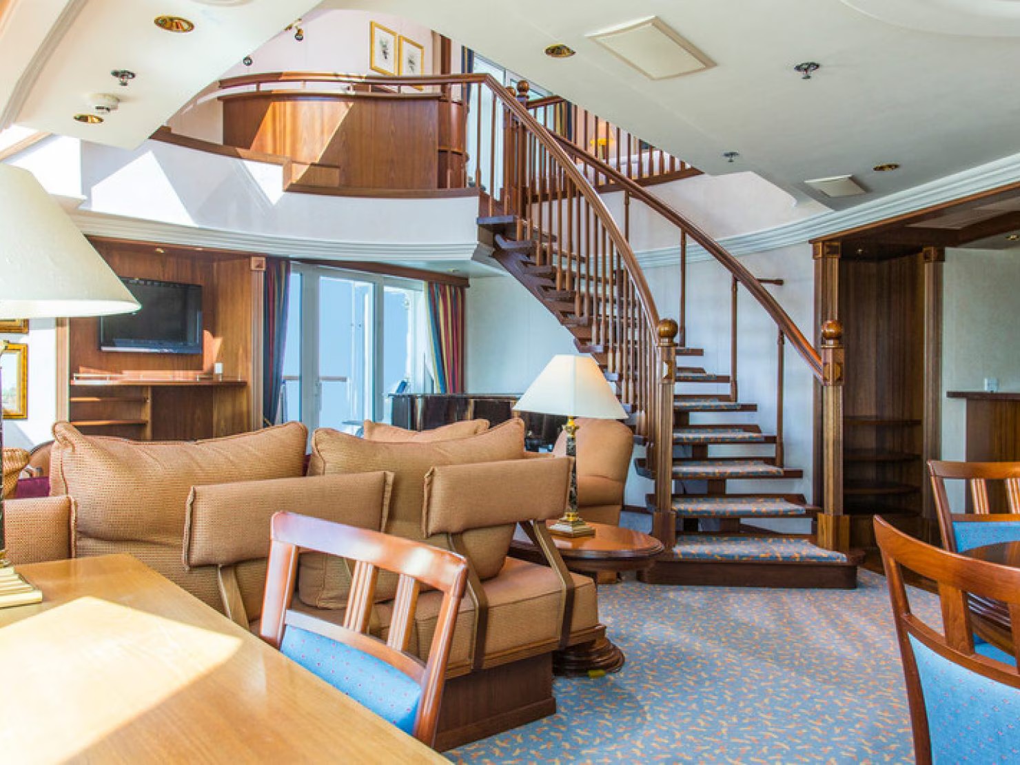 A luxurious penthouse suite aboard a cruise ship featuring a dramatic spiral staircase with wooden railings, elegant furnishings in warm tones, and a two-story open layout.