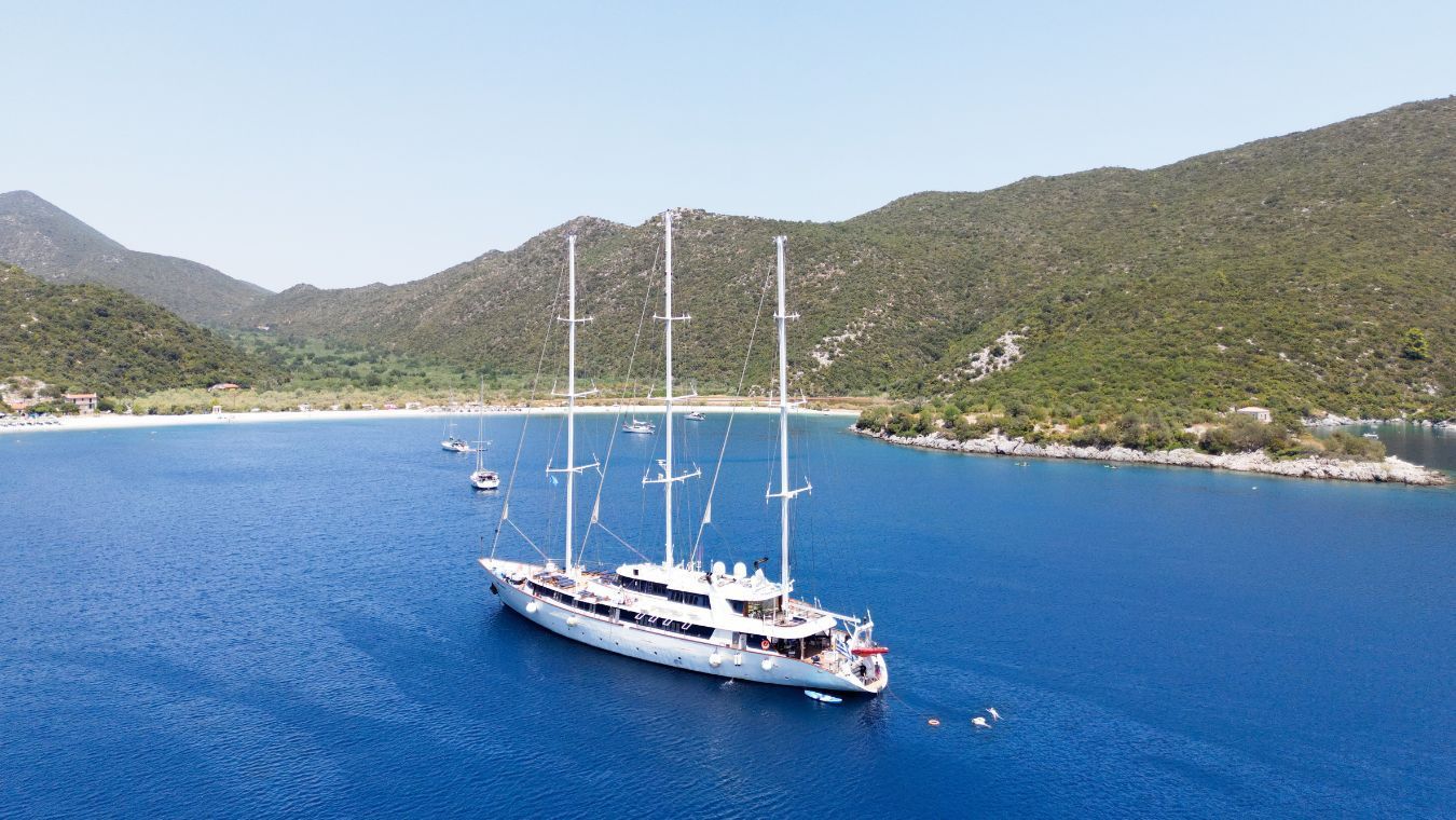 panorama-5-625c233249 A stunning white multi-masted gulet sailing yacht anchors in crystal-clear turquoise waters of a picturesque Mediterranean bay, surrounded by verdant pine-covered mountains and a charming coastal village.