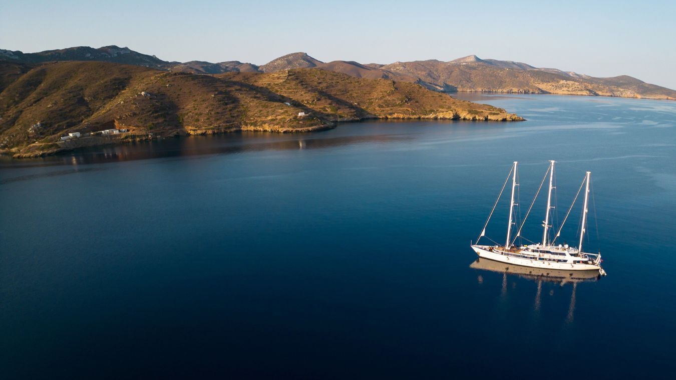 panorama-6-ddf6b47d11 A stunning aerial view of a classic three-masted sailing vessel anchored in crystalline blue waters, with a scenic Mediterranean coastline featuring golden-brown hills and scattered white buildings.