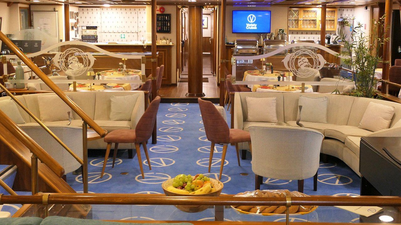 panorama-lounge-1-556c3a79b3 A sophisticated panorama lounge aboard a cruise ship featuring cream-colored seating arranged around dining tables, a blue patterned carpet with geometric designs, wooden architectural elements, and warm ambient lighting.