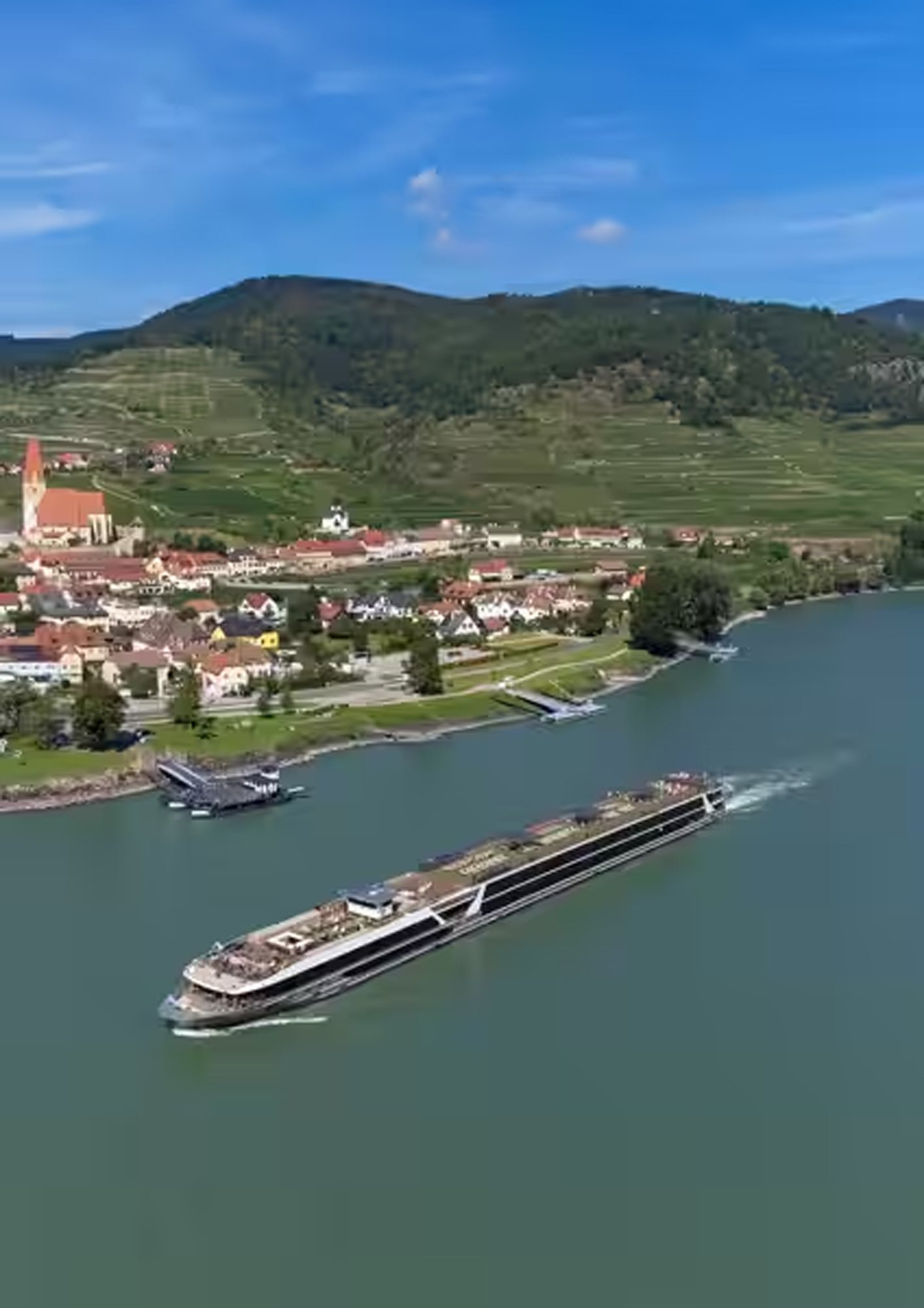 t-eu-vega-river-ship-exterior-03-2022 A modern river cruise ship navigates through a picturesque European river valley, passing a charming riverside town with a distinctive church steeple, terraced vineyards on rolling hills, and traditional architecture.