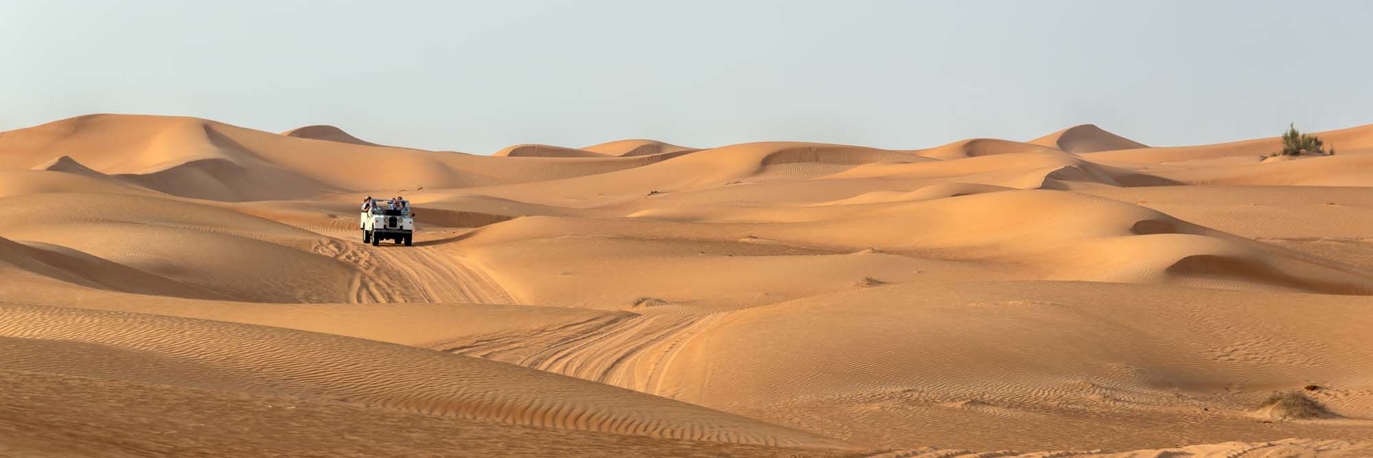 A vintage open-top 4x4 SUV traverses the golden sand dunes of the Dubai desert, navigating the undulating landscape with multiple sand dunes visible under clear skies.