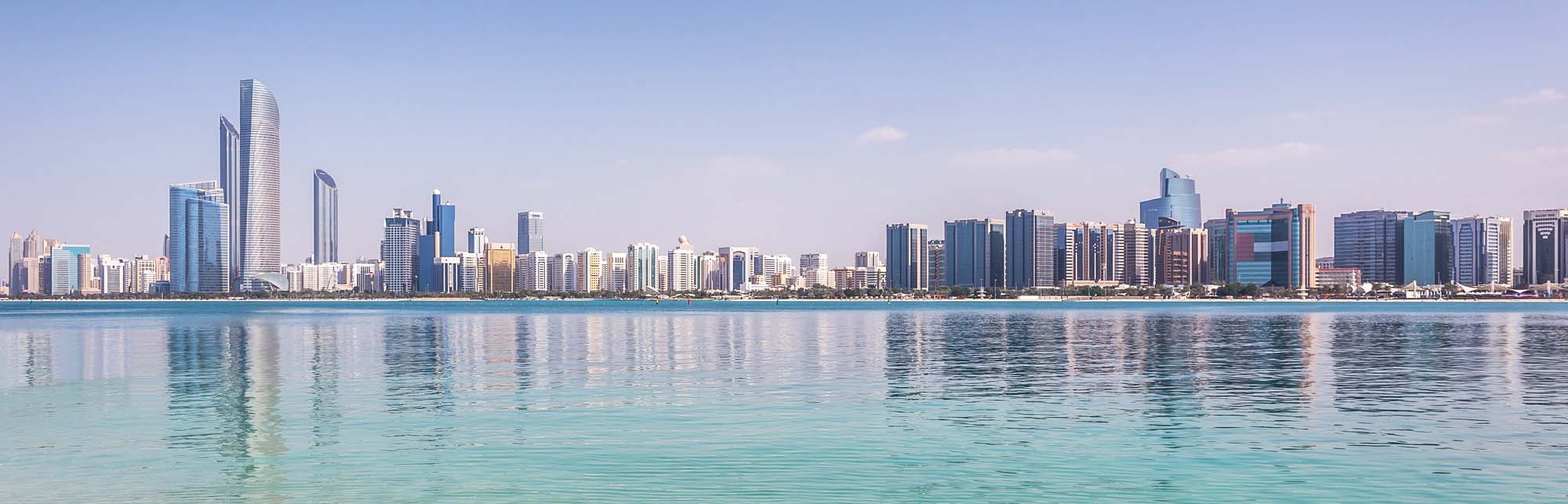 A panoramic view of Abu Dhabi's modern skyline reflecting in calm turquoise waters, showcasing numerous high-rise buildings and skyscrapers along the Persian Gulf coastline under clear blue skies.