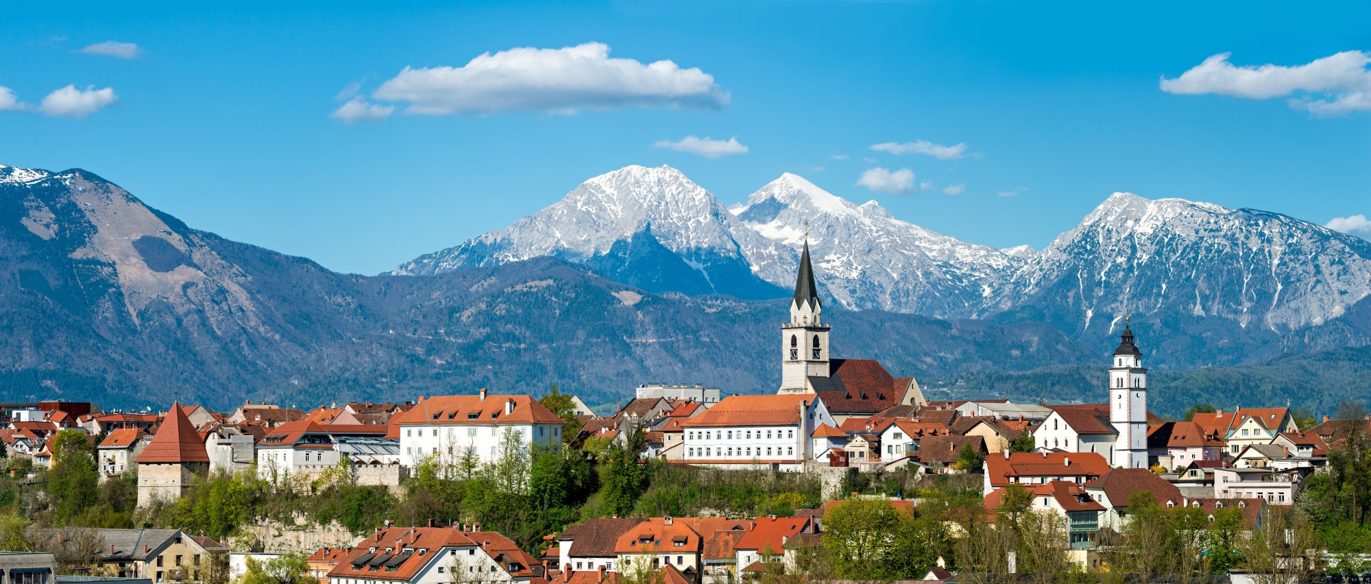A picturesque Alpine village nestled in a valley with distinctive orange-tiled roofs and white church spires, framed by dramatic snow-covered mountain peaks in the background under a clear blue sky.