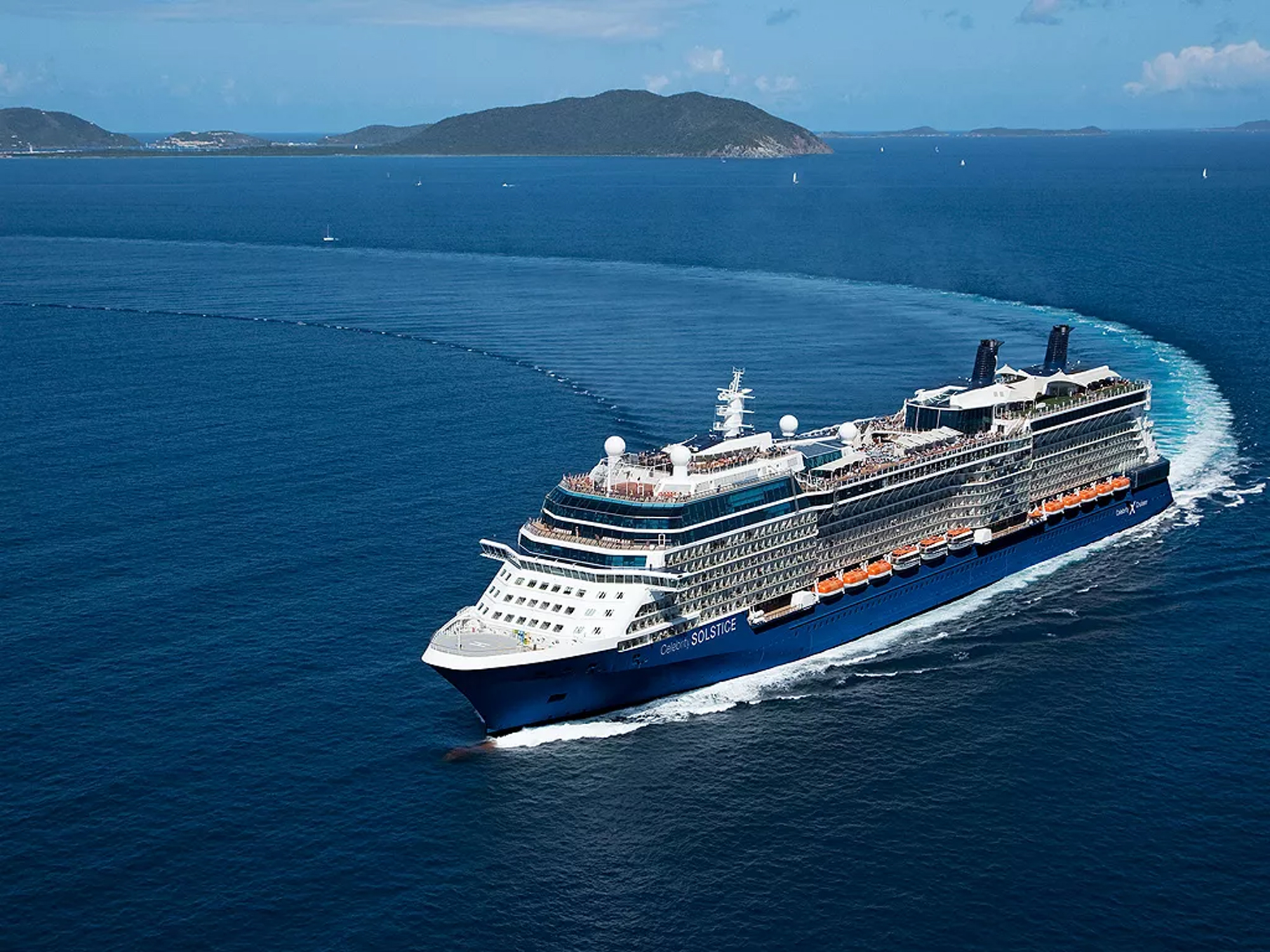A large modern cruise ship with distinctive blue hull and orange lifeboats cruises through deep blue waters near a mountainous island coastline.
