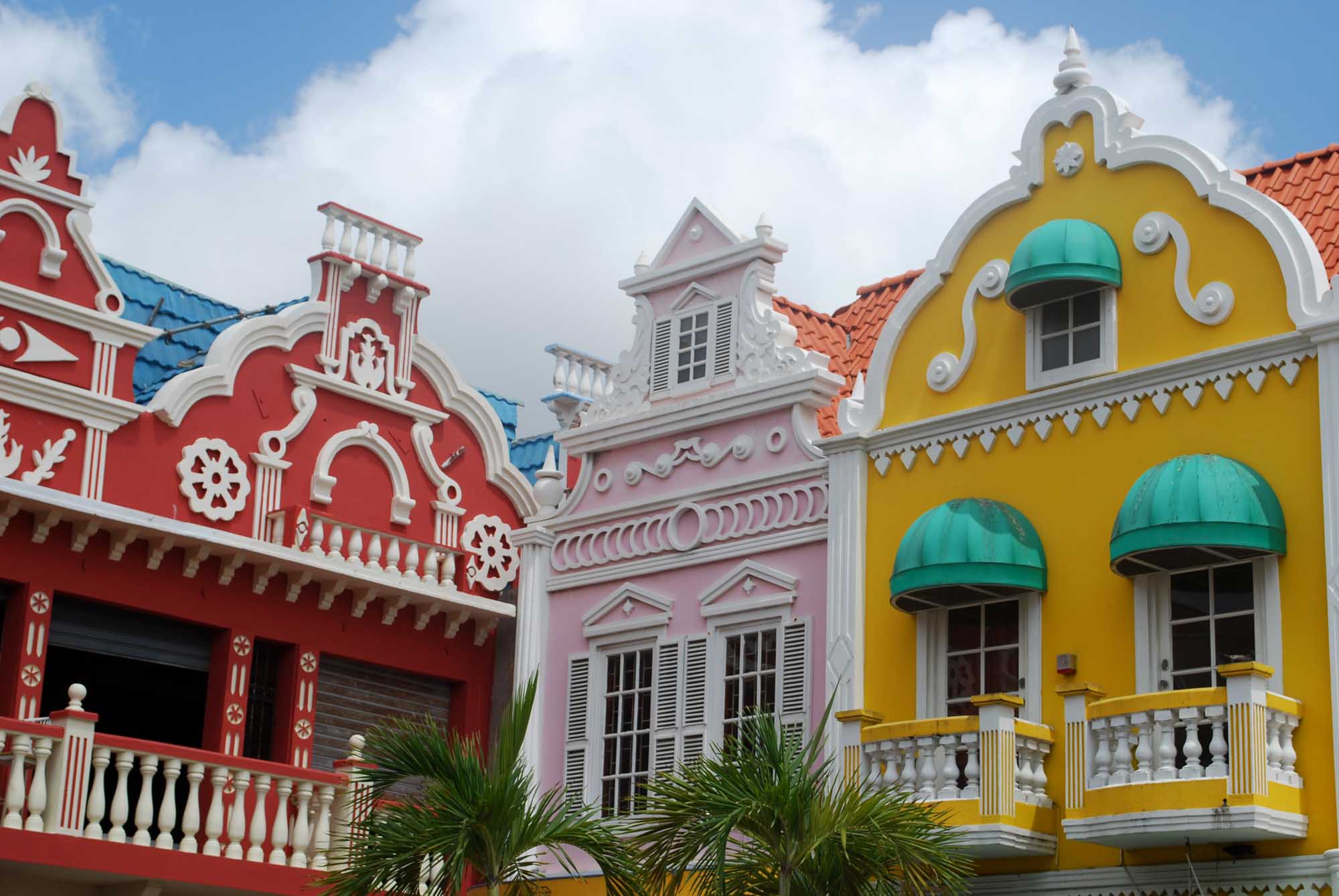 Colorful Colonial Architecture in Willemstad A vibrant row of Dutch Colonial buildings featuring bold primary colors - red, pink, and yellow - with white ornamental trim, arched windows, and turquoise awnings. The iconic architecture reflects Caribbean colonial heritage with distinctive peaked roofs and decorative balustrades, typical of Curacao's historic waterfront district.