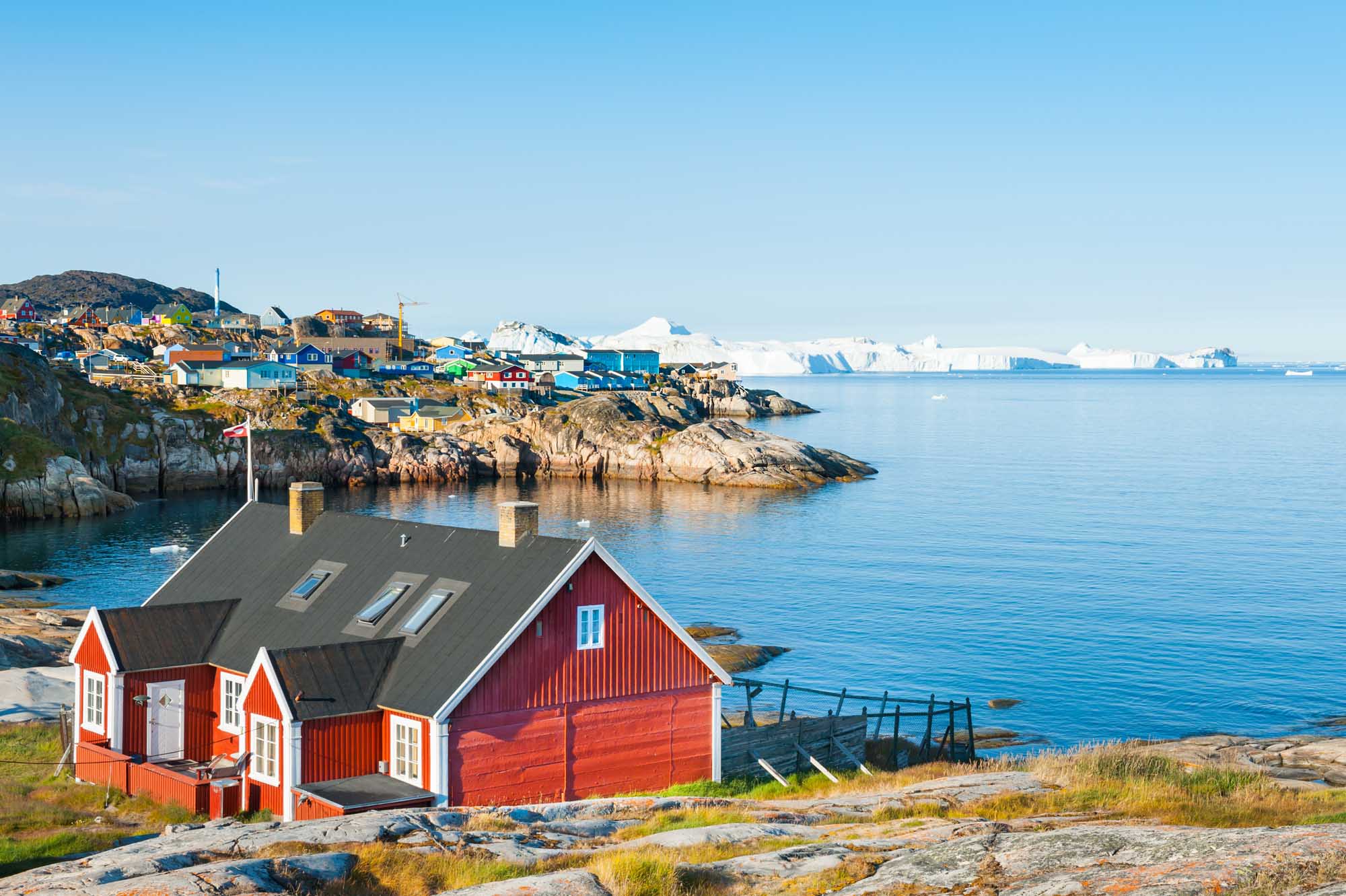 Colorful Houses in Ilulissat, Greenland A picturesque red Greenlandic house sits on rocky terrain overlooking the Atlantic Ocean, with the vibrant settlement of Ilulissat visible across the bay and icebergs dotting the horizon.
