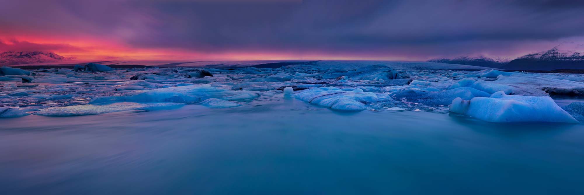 A stunning twilight panorama of Iceland's Jokulsarlon glacier lagoon, featuring icebergs floating in crystalline turquoise water beneath a dramatic sunset sky painted in shades of crimson, purple, and deep blue.
