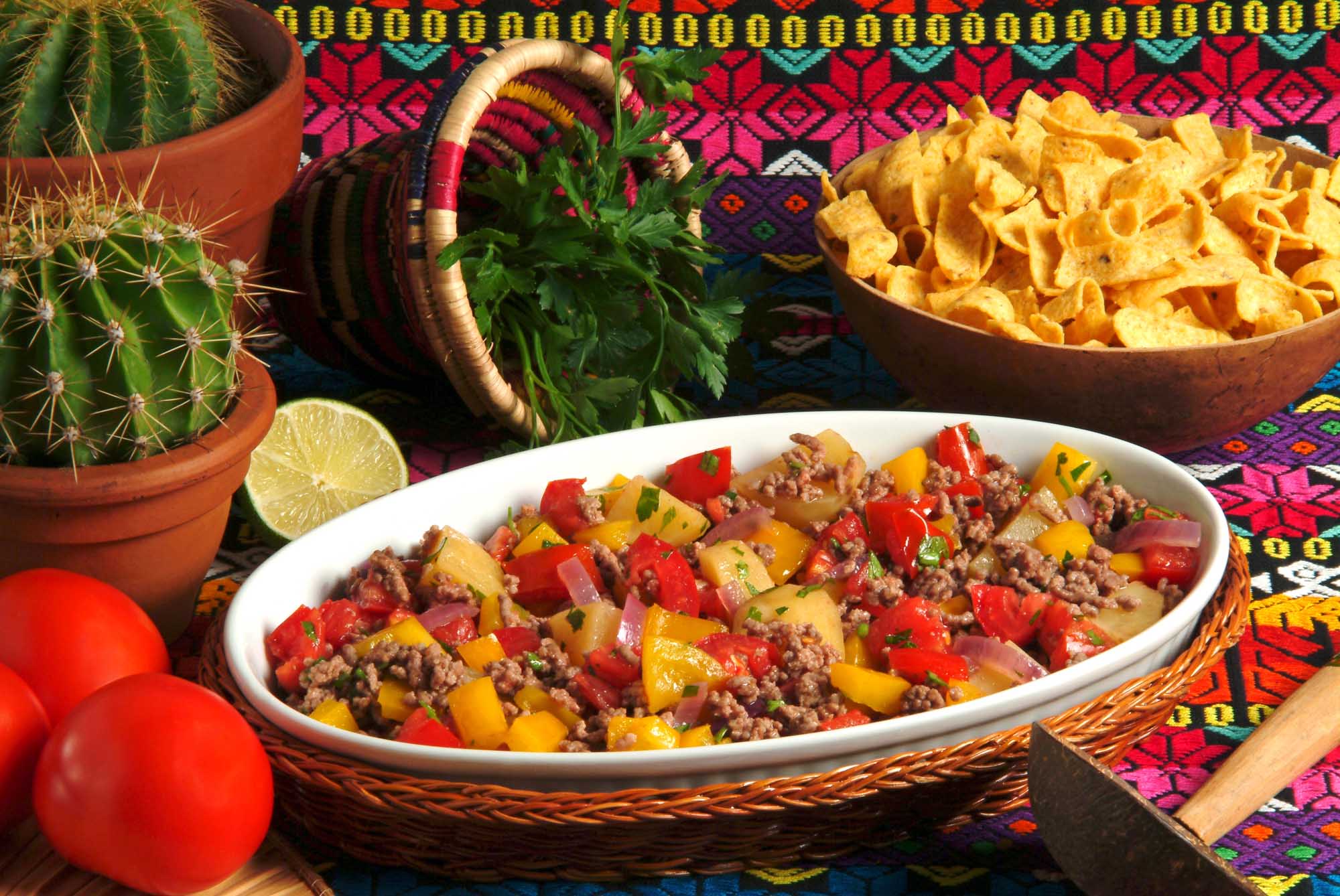 Cuban Picadillo with Traditional Styling A vibrant presentation of Cuban picadillo habanero served in a white ceramic bowl, featuring seasoned ground meat with diced potatoes, red and yellow peppers, and fresh cilantro garnish. The dish is artfully arranged on a colorful woven placemat alongside complementary items including fresh lime, a decorative ceramic pitcher, potted cactus, crispy tortilla chips, and fresh tomatoes, creating an authentic and appetizing Cuban culinary scene.
