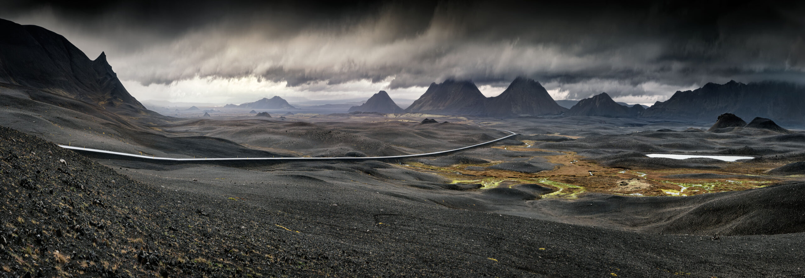 A sweeping panoramic view of Iceland's stark volcanic highlands, featuring a winding glacial river cutting through dark lava fields, dramatic jagged mountain peaks in the distance, and moody storm clouds overhead.