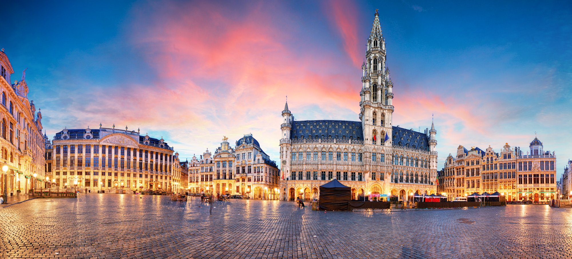 A stunning twilight view of Brussels' iconic Grand Place (Grote Markt) featuring the ornate Gothic Town Hall with its distinctive spire, surrounded by elegant 17th-century guild houses illuminated by warm golden lights against a vibrant blue and pink sunset sky.