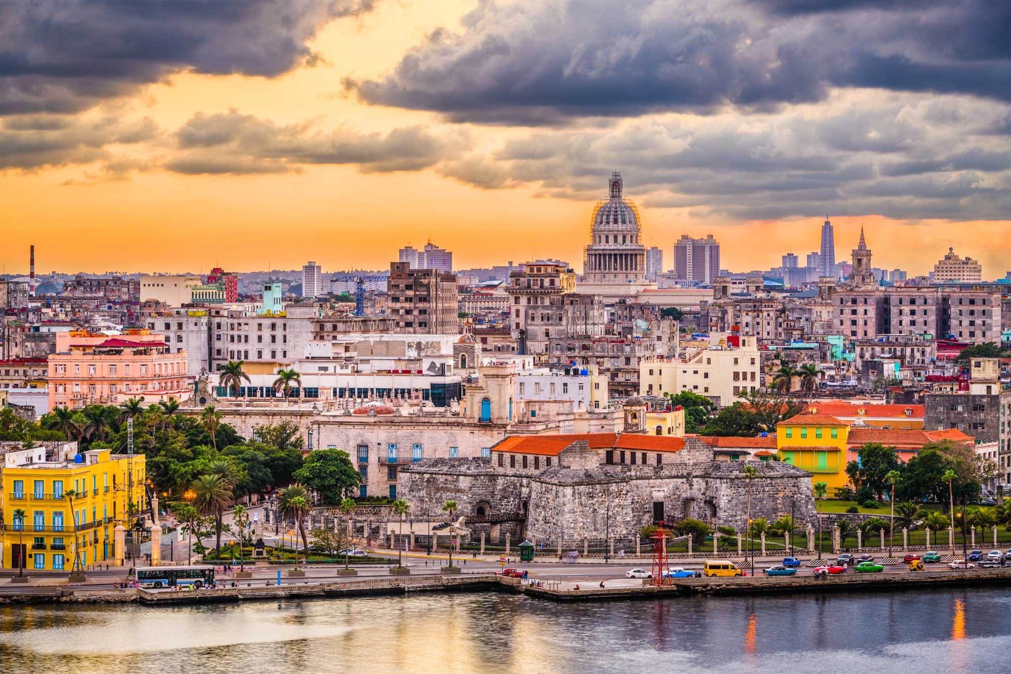 Havana Skyline at Sunset with Capitol Dome A stunning panoramic view of Havana, Cuba's downtown skyline at dusk, featuring the iconic Capitol building with its distinctive dome, colorful colonial architecture, palm trees, and the waterfront promenade (Malecon) along the harbor. The dramatic golden sky with storm clouds creates a striking backdrop for this vibrant Caribbean city.