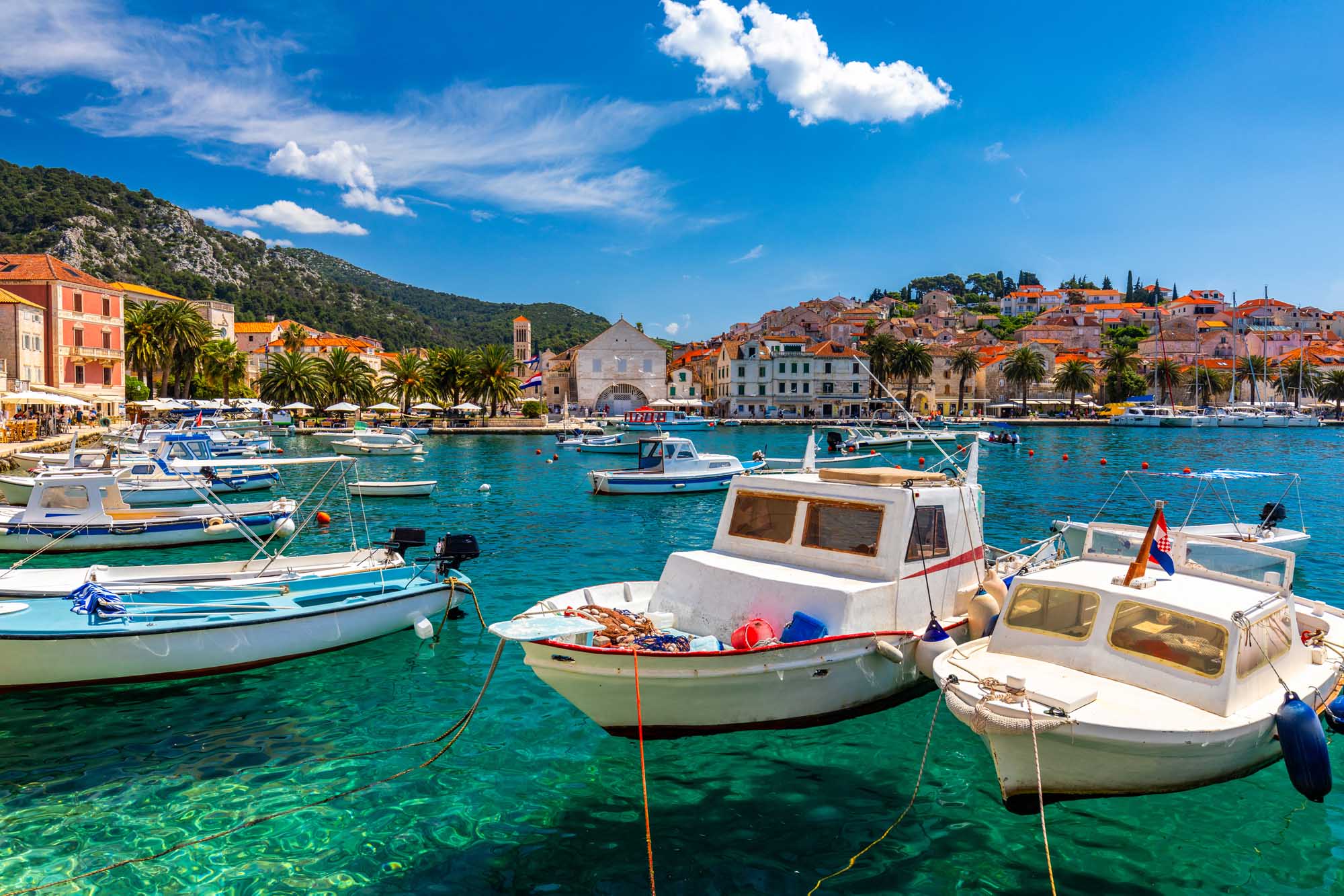A picturesque Mediterranean harbor scene featuring numerous fishing boats anchored in crystal-clear turquoise waters off the coast of Hvar, Croatia. The historic town rises dramatically behind the harbor with traditional orange-roofed buildings nestled against a verdant hillside.