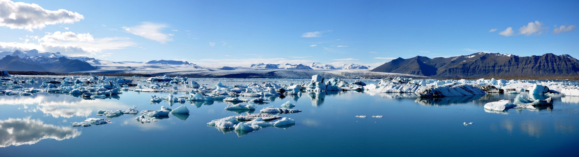 A breathtaking panoramic view of Jokulsarlon Glacier Lagoon in Iceland, featuring countless blue and white icebergs floating in pristine turquoise waters. Snow-capped mountains frame the horizon under a clear blue sky with scattered white clouds.