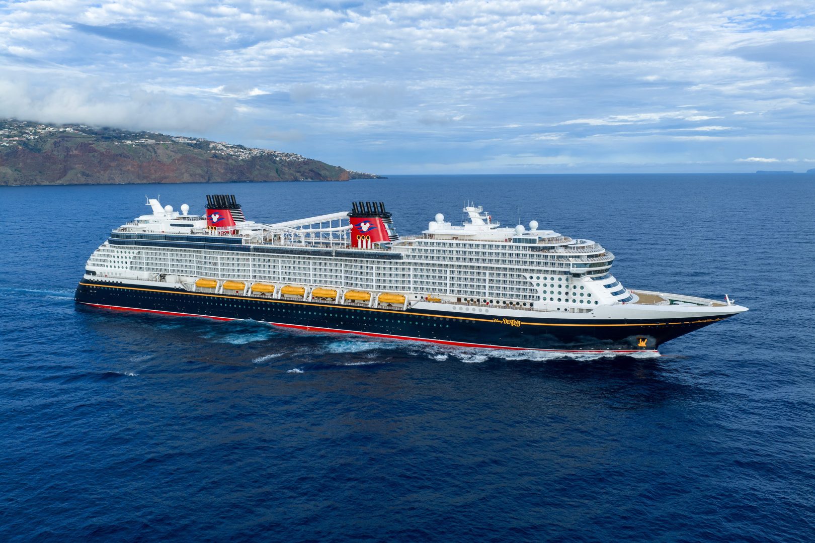 Large Cruise Ship Anchored Off Volcanic Island A massive modern cruise ship with distinctive red funnels featuring Mickey Mouse imagery is anchored in brilliant blue waters off a volcanic island with terraced settlements.