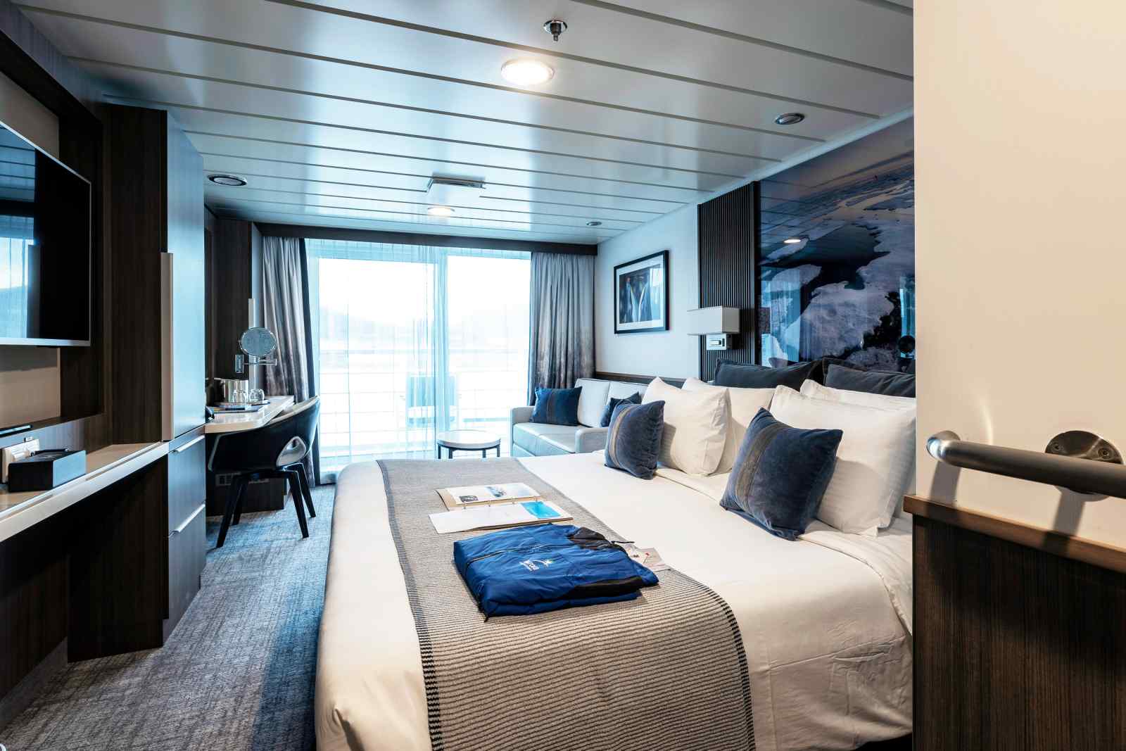 Luxury Cruise Ship Stateroom with Ocean View A luxurious cruise ship stateroom featuring a king-size bed with premium bedding, floor-to-ceiling windows with ocean views, modern furnishings in dark wood tones, a work desk, and sophisticated blue and white color scheme accents throughout the elegantly appointed cabin.