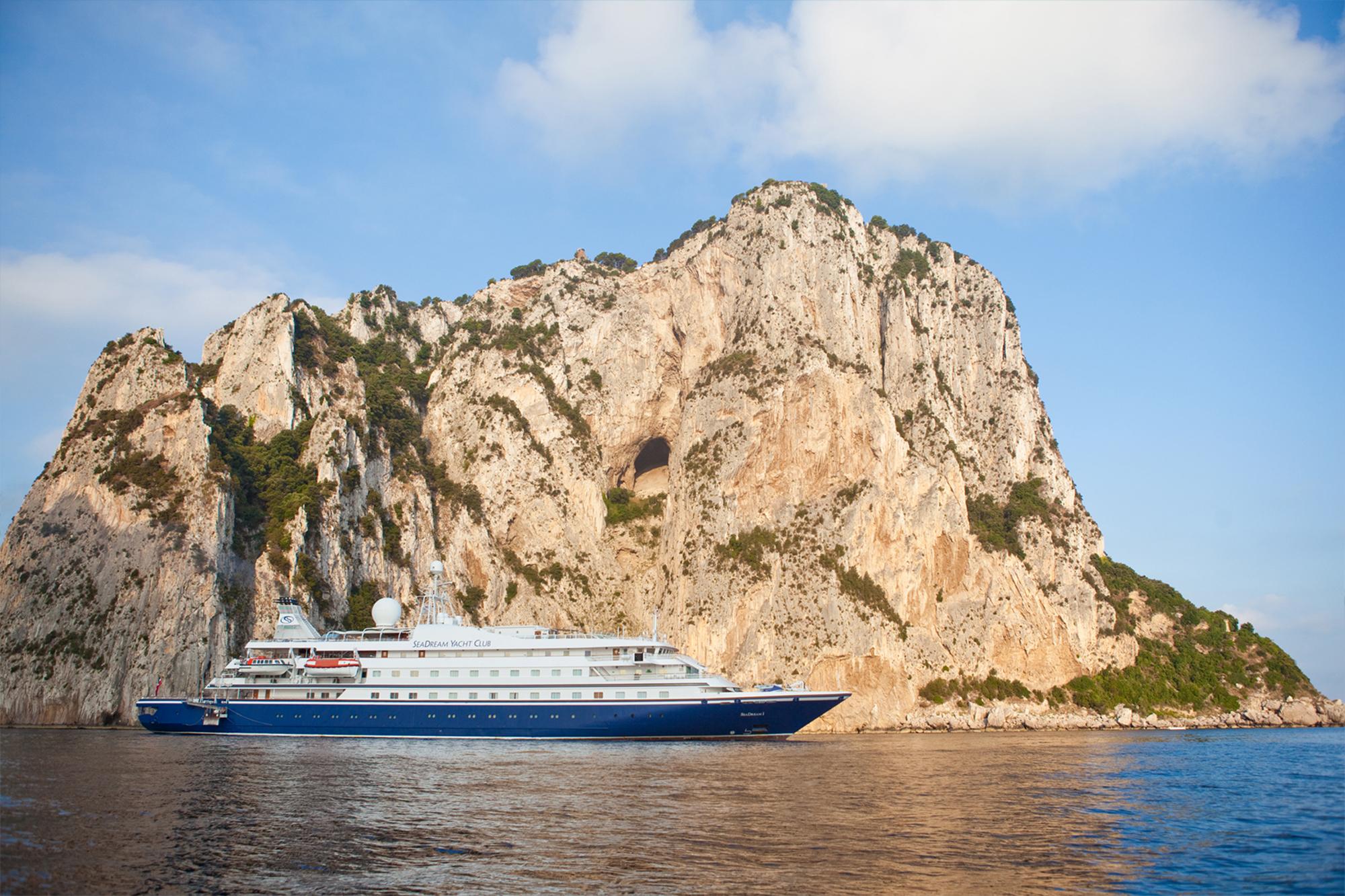 A large multi-deck cruise ship anchors in the azure waters off Capri, Italy, positioned against the dramatic limestone cliffs and natural rock formations of the Faraglioni.