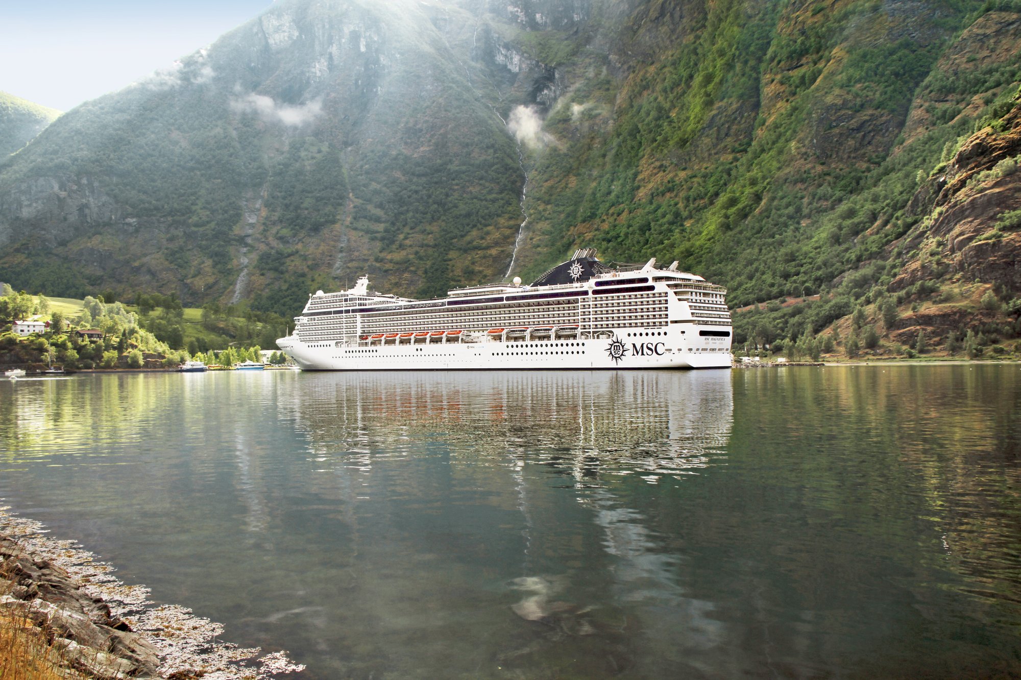A large MSC cruise ship navigates through a dramatic Norwegian fjord surrounded by steep, forested mountain cliffs and cascading waterfalls.