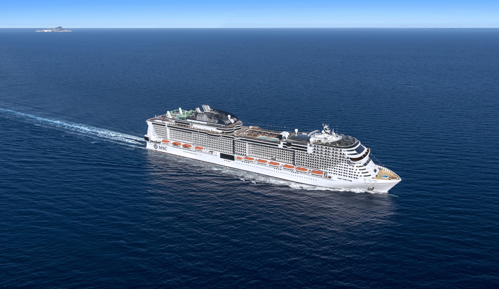 A modern mega-ship cruise liner traveling through clear blue Mediterranean waters under bright sunshine. The vessel displays distinctive MSC branding and orange accent striping along its white hull, with multiple decks visible and a small island visible in the distant horizon.