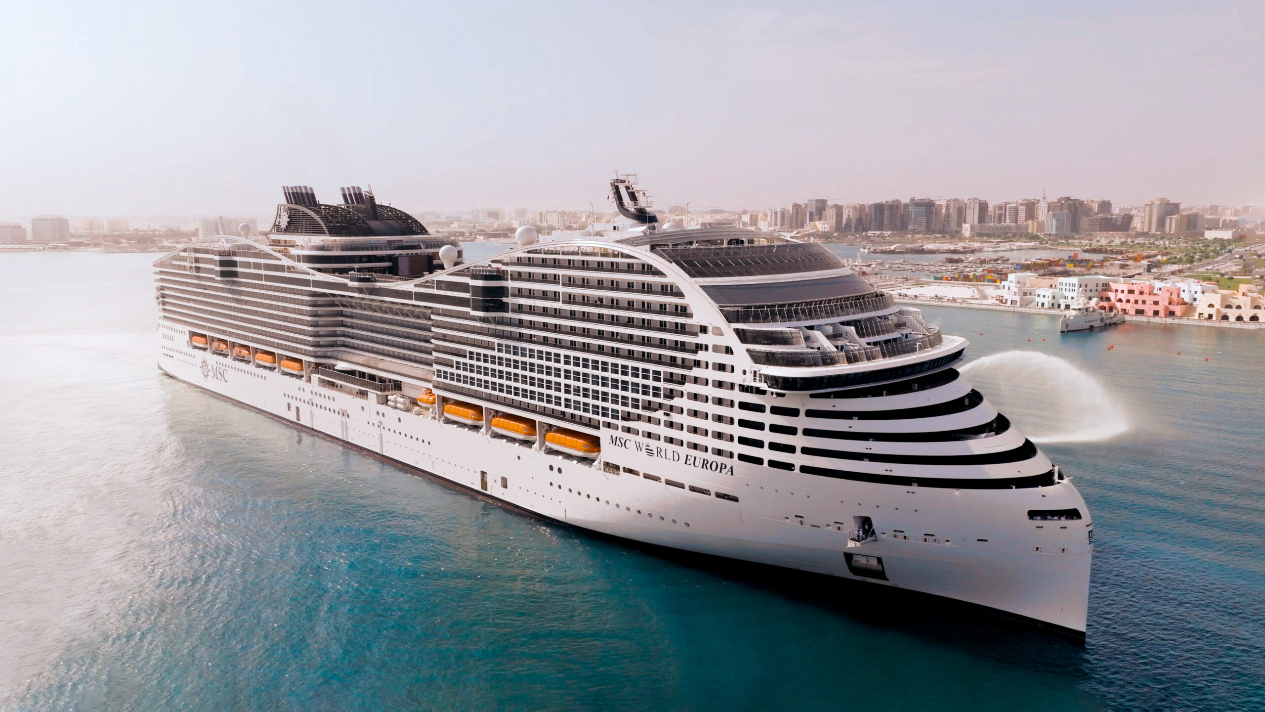 A large modern cruise ship, the MSC World Europa, is anchored in turquoise waters near a Mediterranean coastal city with white and coral-colored waterfront buildings visible in the background.