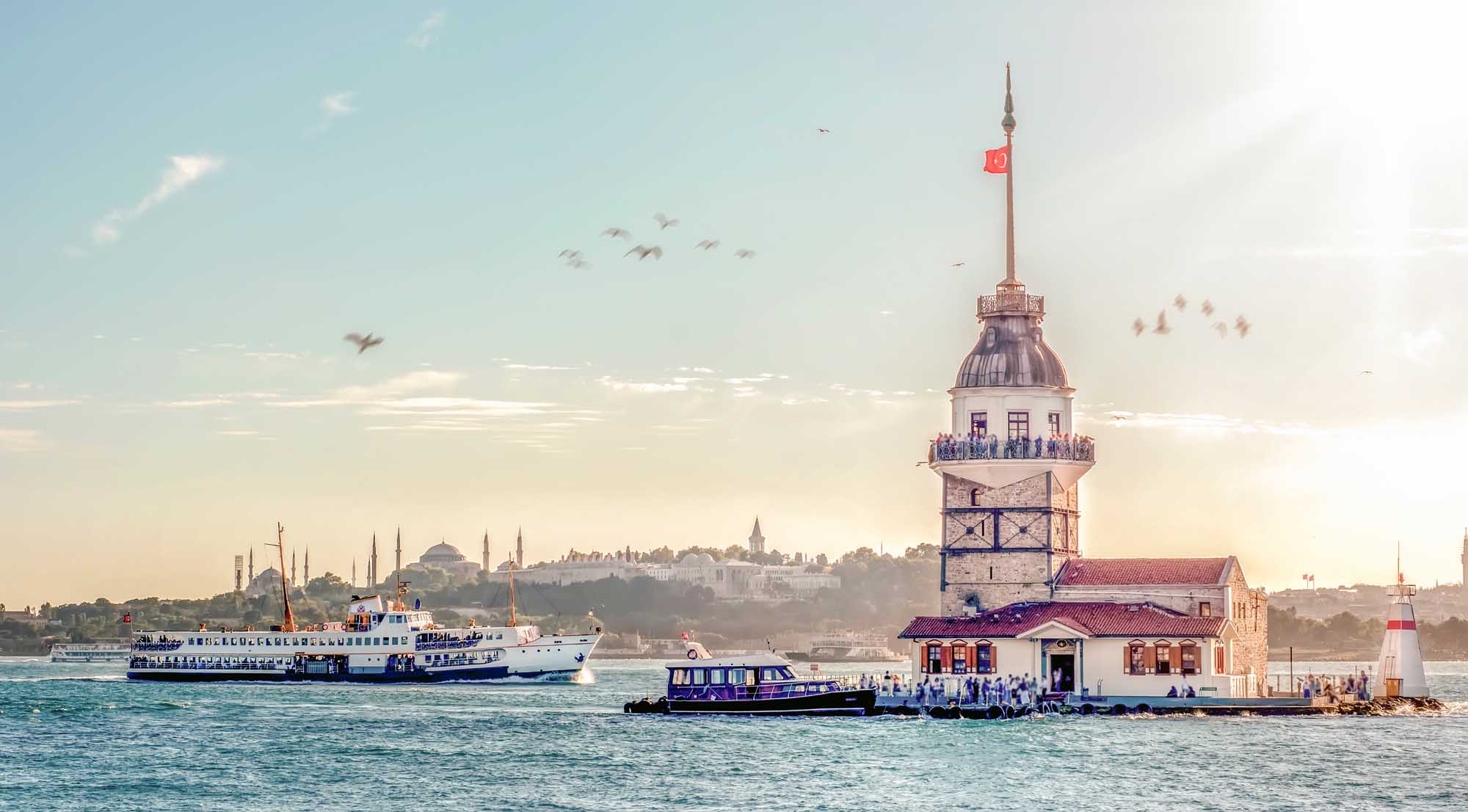 Maiden’s Tower Istanbul with Ferry Boats A scenic view of Istanbul's iconic Maiden's Tower (Kiz Kulesi) in the Bosphorus Strait, with a blue and white passenger ferry approaching the tower and the cityscape of Istanbul visible in the background under a clear sky.