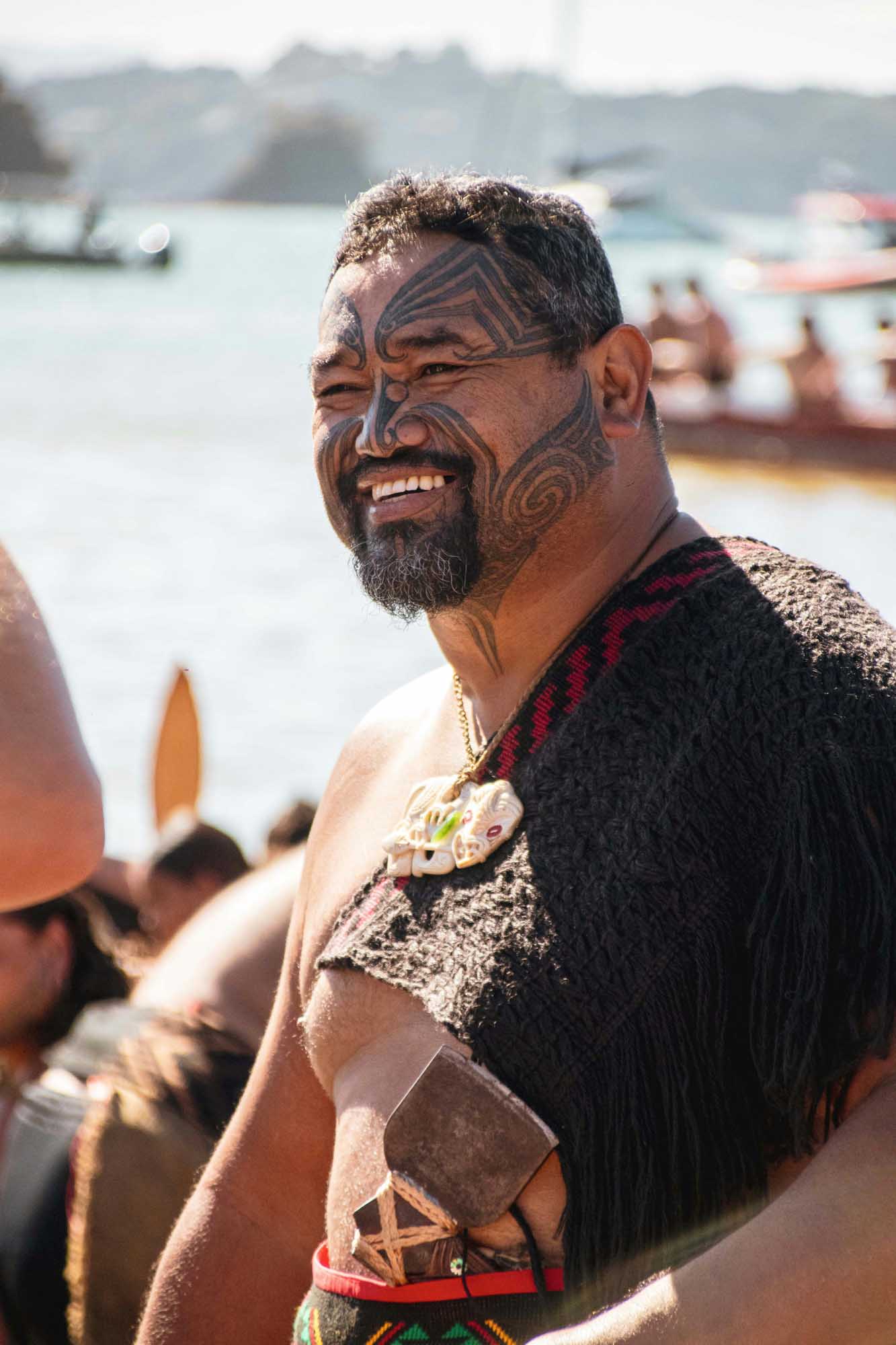 Maori Man with Traditional Facial Tattoo A man with traditional Maori ta moko facial tattoos smiles at the camera while standing near a waterfront with boats and hills in the background.