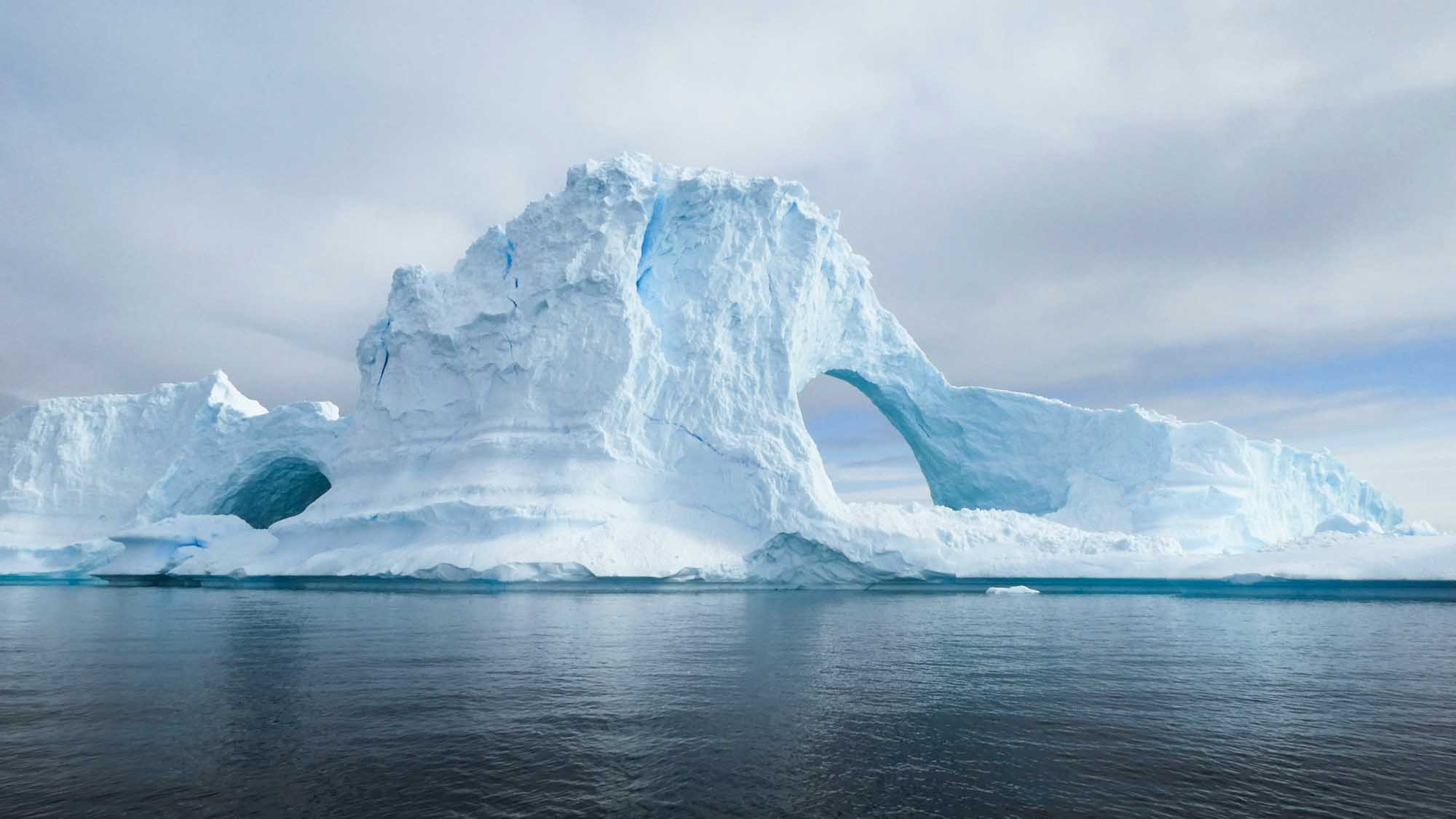 Massive Iceberg with Natural Arch Formation A spectacular tabular iceberg featuring a distinctive natural arch formation carved through its center, floating in calm turquoise waters.