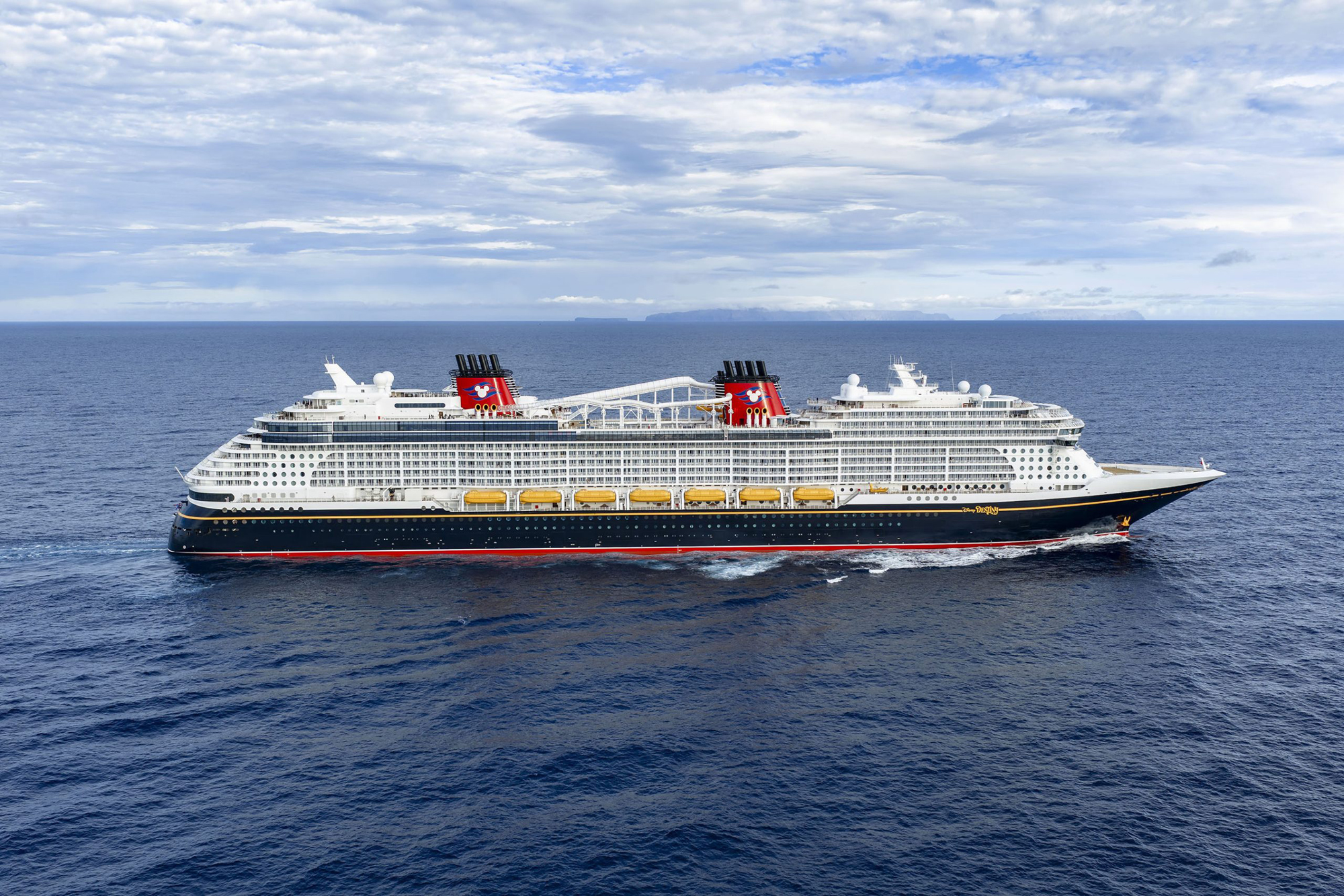 Modern Cruise Ship at Sea with Mountain Vista A large modern cruise ship with distinctive red funnels sails through calm blue waters under a partly cloudy sky with mountainous terrain visible on the horizon.