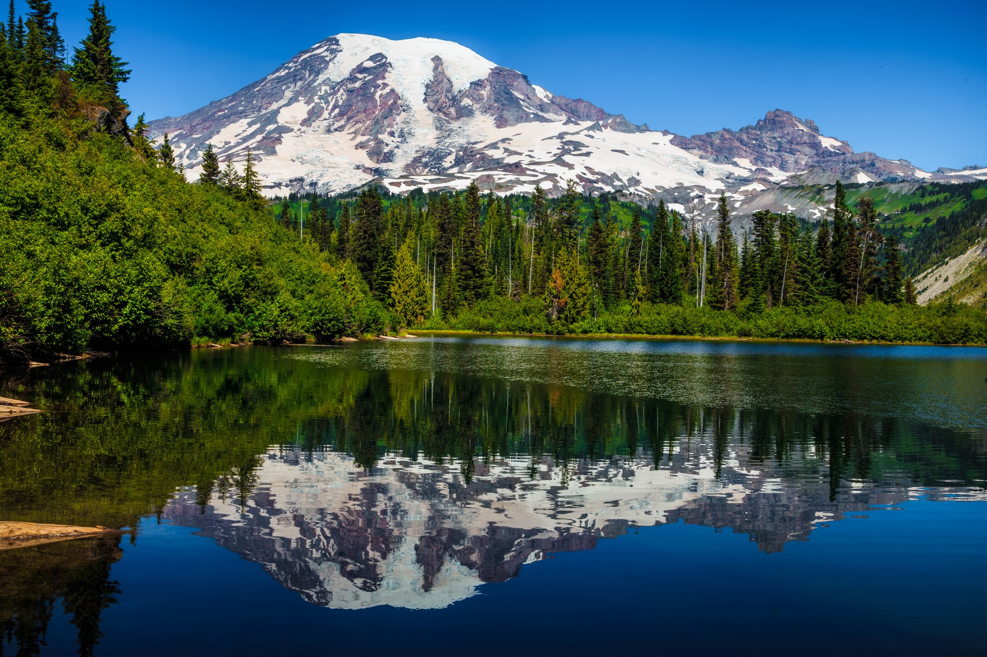 Mount Rainier Reflection in Alpine Lake A stunning alpine landscape featuring Mount Rainier's snow-capped summit perfectly reflected in a pristine mountain lake, surrounded by dense coniferous forest under clear blue skies.