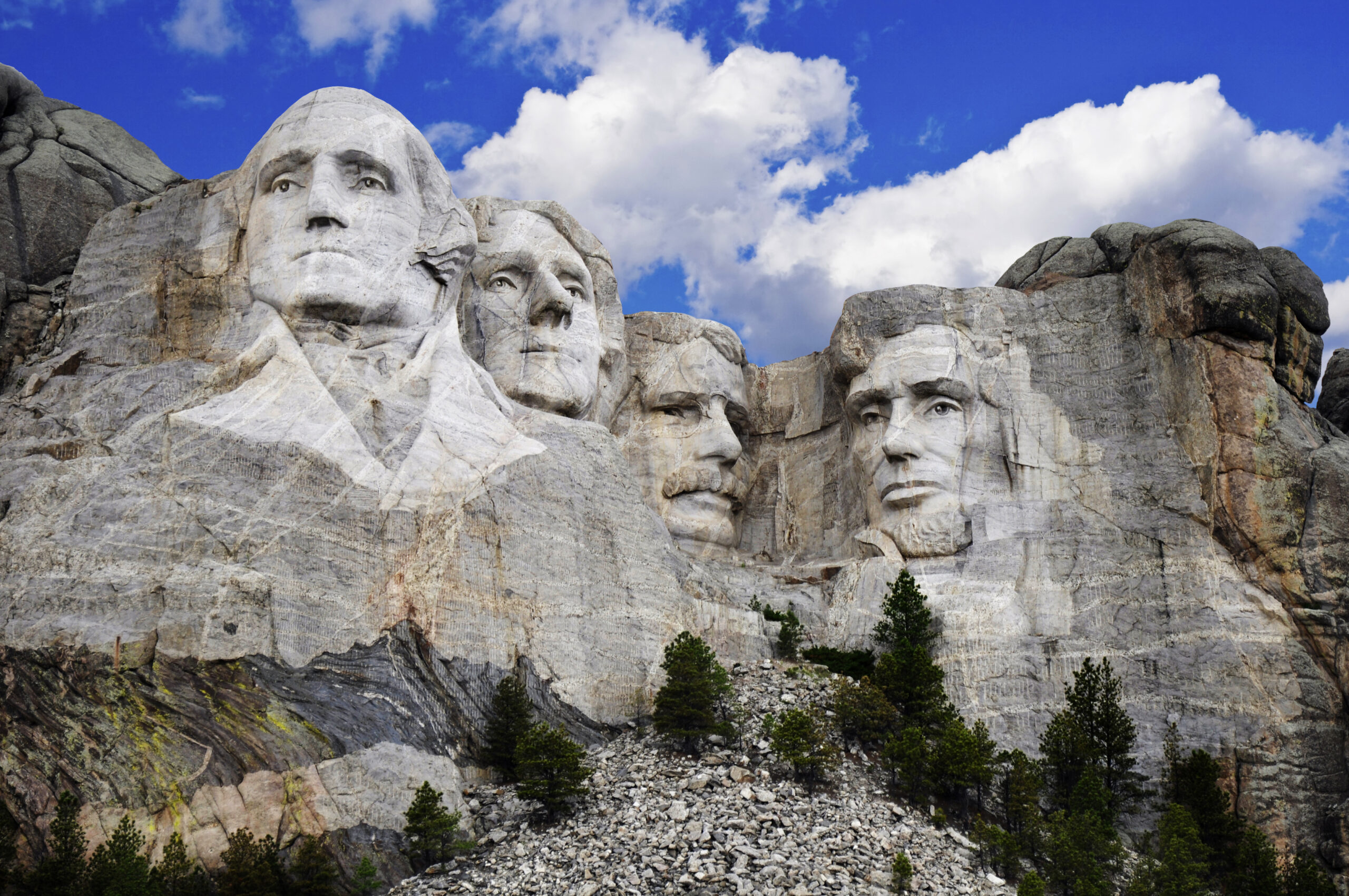 Mount Rushmore National Monument South Dakota The iconic Mount Rushmore monument featuring four carved presidential faces (Washington, Jefferson, Theodore Roosevelt, and Lincoln) carved into granite cliffs against a brilliant blue sky with white clouds. Evergreen trees dot the base of the mountain.