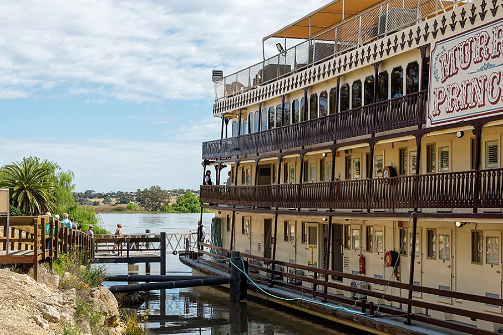A three-deck paddlewheel riverboat with cream and brown colonial-style architecture docked along a peaceful river, featuring ornate railings and multiple levels of verandahs.