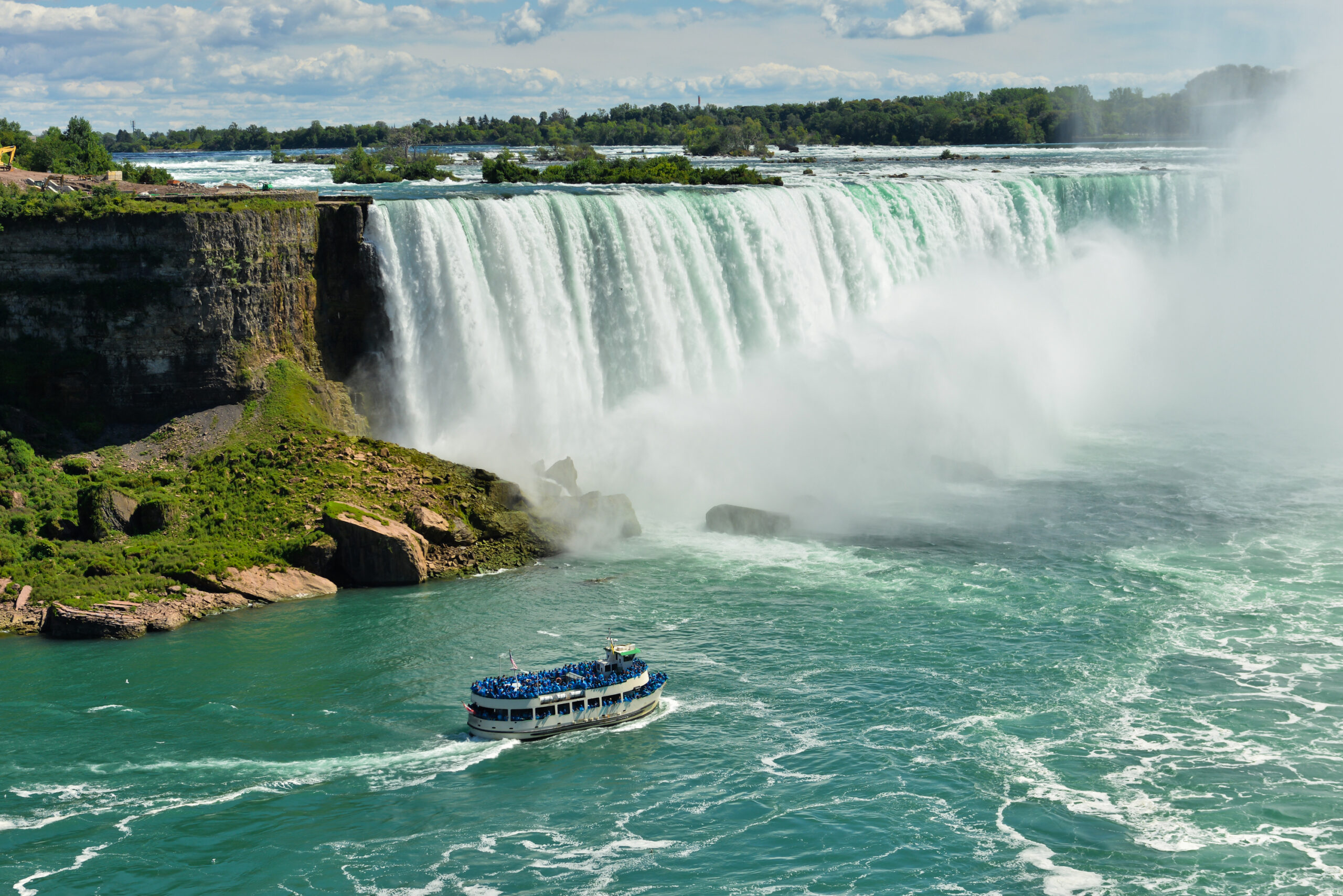 Niagara Falls with Tourist Boat Tour A scenic view of Niagara Falls Horseshoe Falls with a passenger tour boat navigating the turquoise waters below, surrounded by mist and lush greenery on the Canadian side. The iconic natural wonder showcases the massive cascade of water with a blue-sky backdrop and forested landscape.