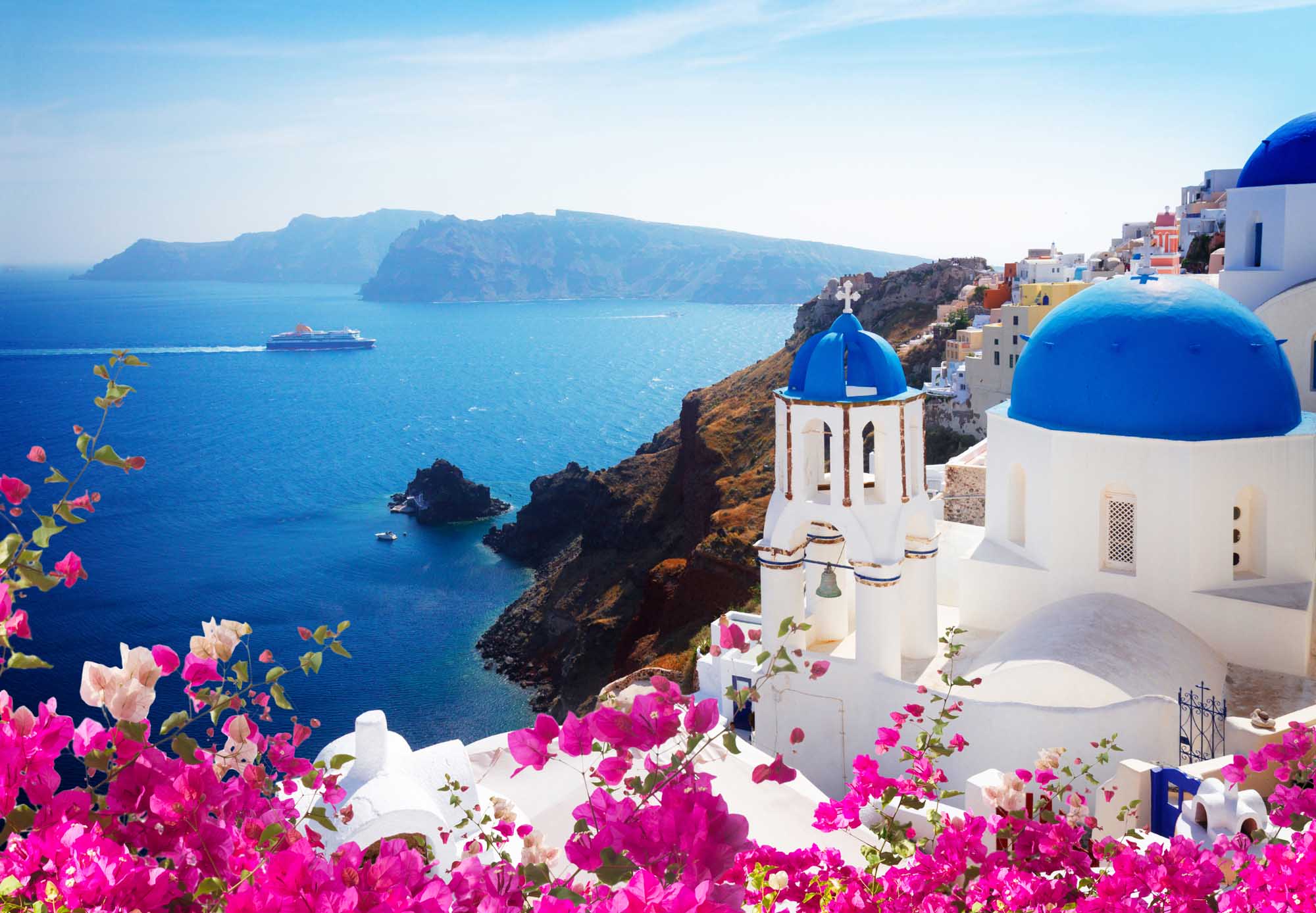 A picturesque view of Oia village in Santorini, Greece, featuring iconic white-washed buildings with brilliant blue-domed churches overlooking the deep blue Aegean Sea with a cruise ship visible in the distance.