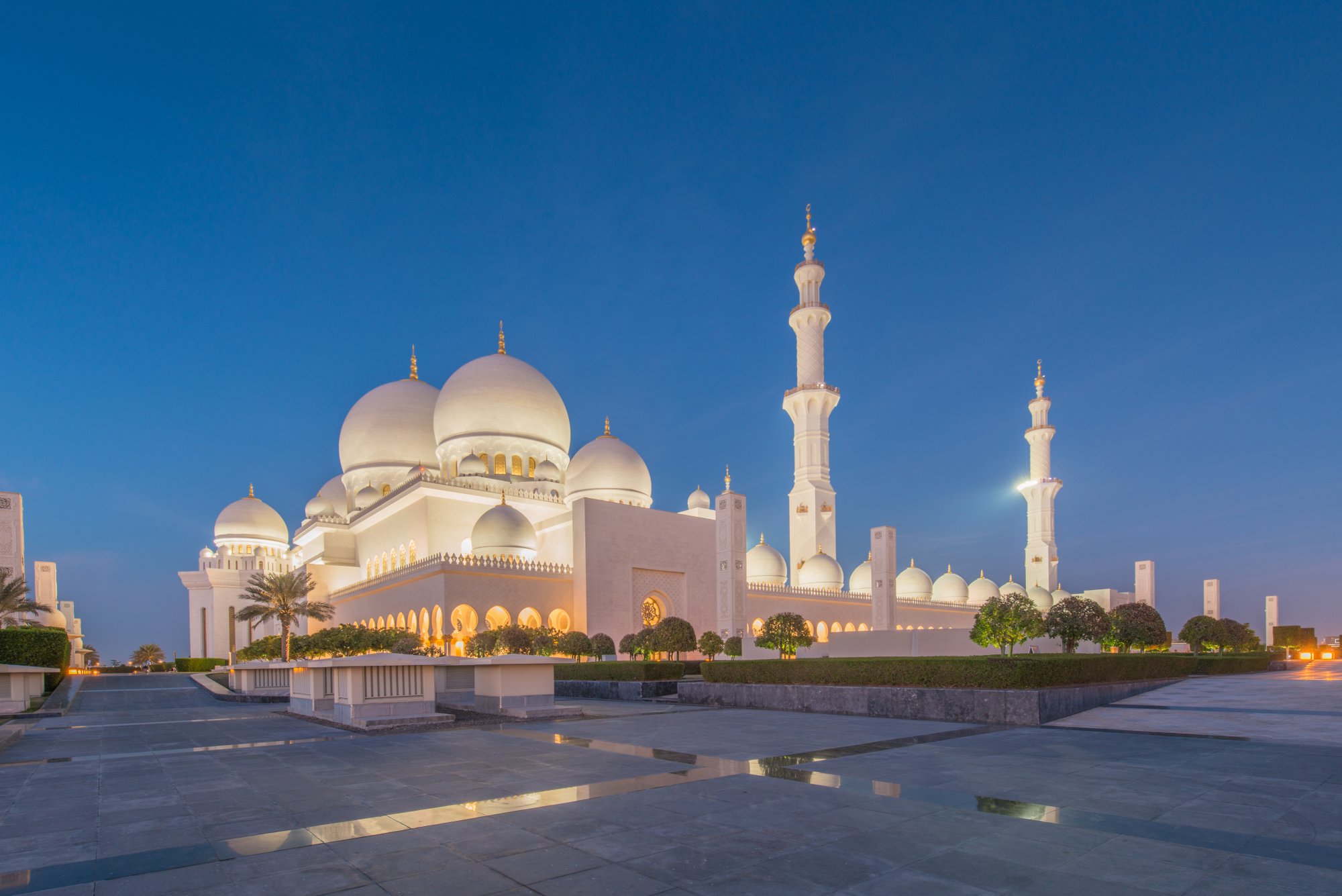 The iconic Sheikh Zayed Grand Mosque illuminated at dusk against a deep blue sky, featuring pristine white marble architecture with multiple domes and minarets, reflected in the still water of the courtyard plaza.