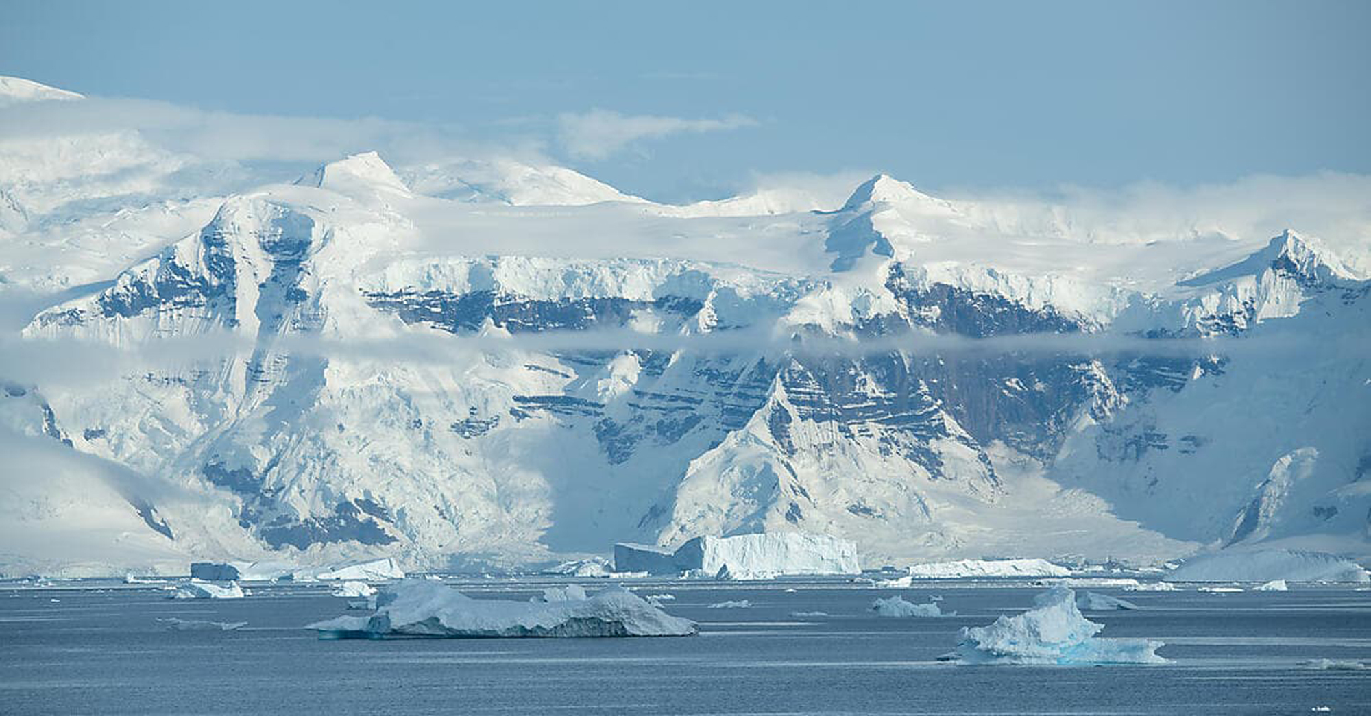 A stunning polar landscape featuring towering snow-covered mountains rising dramatically from the sea, with numerous icebergs floating in the foreground. The pristine white peaks are partially shrouded in mist, creating a serene and majestic Antarctic seascape under clear blue skies.