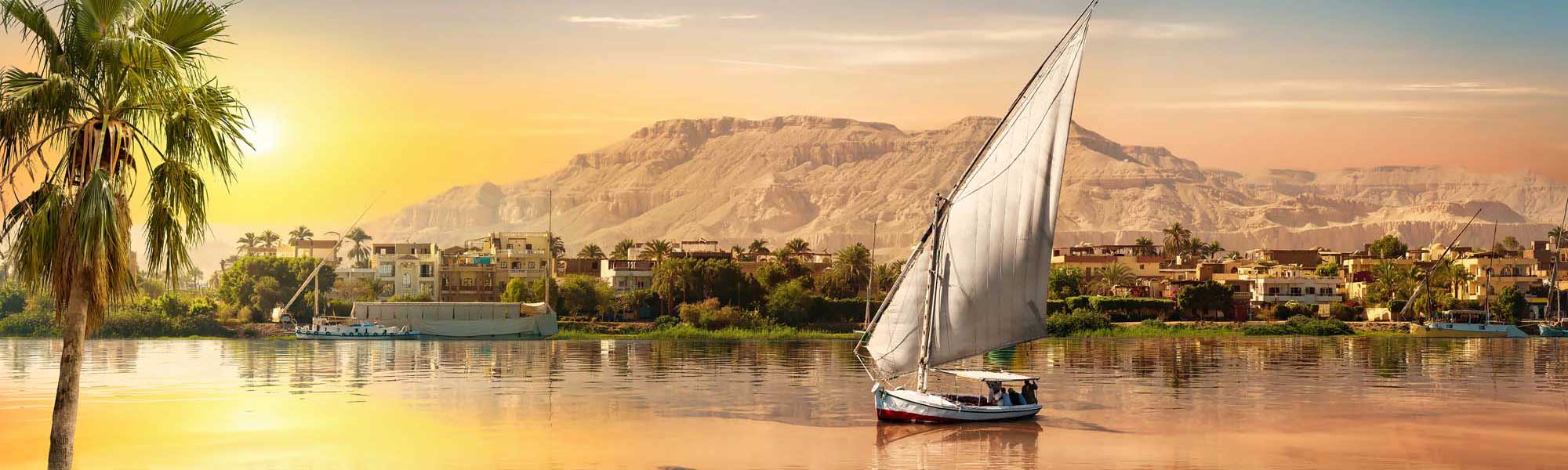 Traditional Felucca Sailing on the Nile River A traditional Egyptian felucca sailboat with white canvas sails glides peacefully across the Nile River at golden sunrise, with palm-lined riverbanks, colorful buildings, and desert cliffs in the background.