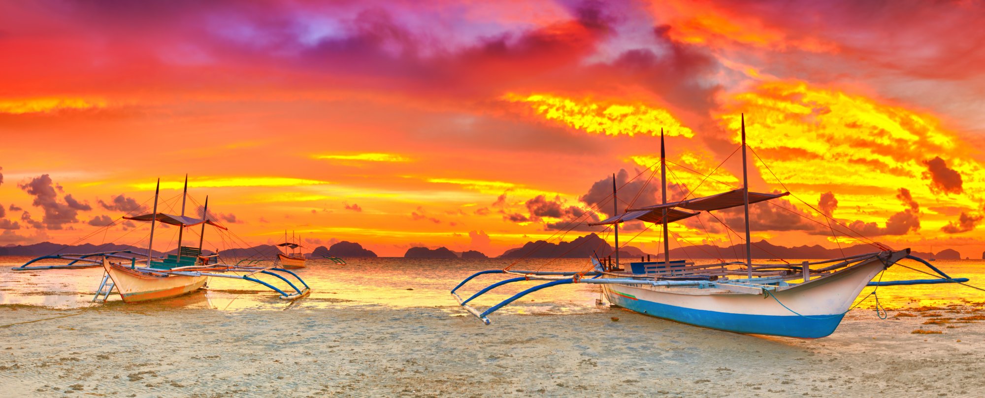 Traditional Outrigger Boats at Tropical Sunset A picturesque tropical beach scene at golden hour featuring traditional Filipino outrigger boats (bancas) with distinctive blue and yellow hulls anchored in shallow waters. The dramatic sunset sky displays vibrant orange, yellow, and purple hues reflected on the calm sea, with distant limestone karsts silhouetted on the horizon, characteristic of Southeast Asian island destinations.