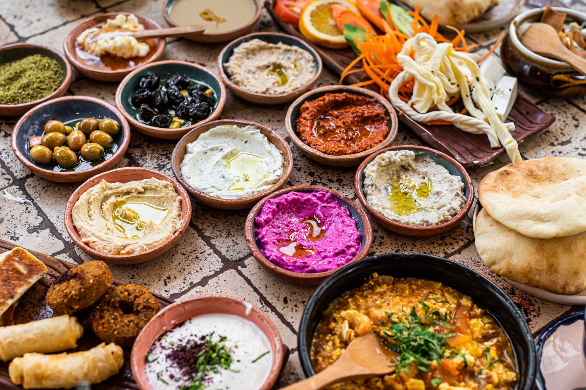 A vibrant overhead spread of authentic Turkish breakfast featuring various mezze dips including hummus, baba ganoush, and muhammara in terracotta bowls, accompanied by fresh vegetables, olives, pastries, bread, and traditional accompaniments artfully arranged on a rustic surface.