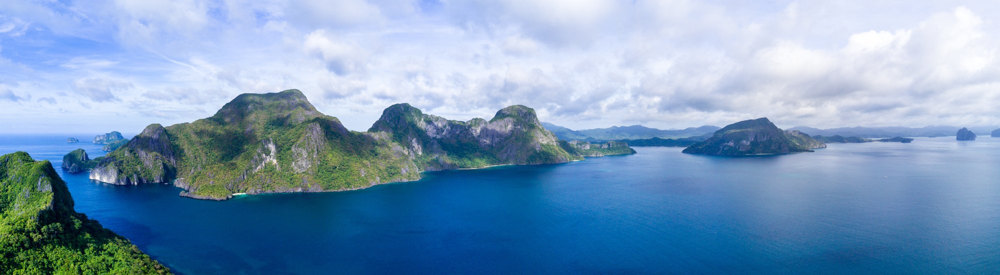 Tropical Island Archipelago in Blue Waters A stunning aerial view of a lush tropical archipelago featuring steep limestone cliffs covered in dense green vegetation, rising dramatically from crystal-clear turquoise waters. Multiple islands of varying sizes are scattered across the seascape under a partly cloudy sky, suggesting a Southeast Asian destination popular with cruise itineraries.