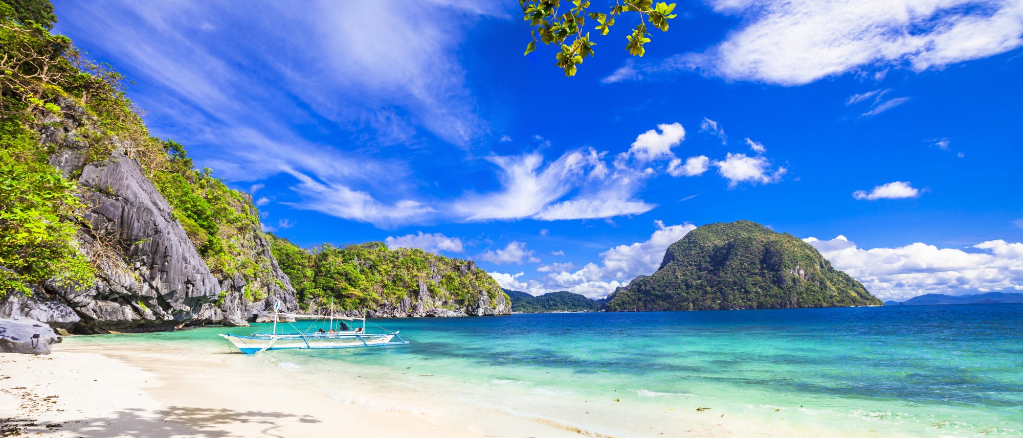Tropical Island Beach Cove with Traditional Boats A pristine tropical beach nestled beneath towering limestone cliffs, featuring crystal-clear turquoise waters, white sand, and traditional outrigger boats anchored offshore. Lush green vegetation covers the dramatic rock formations, with a distant volcanic island visible across the bay under a bright blue sky.