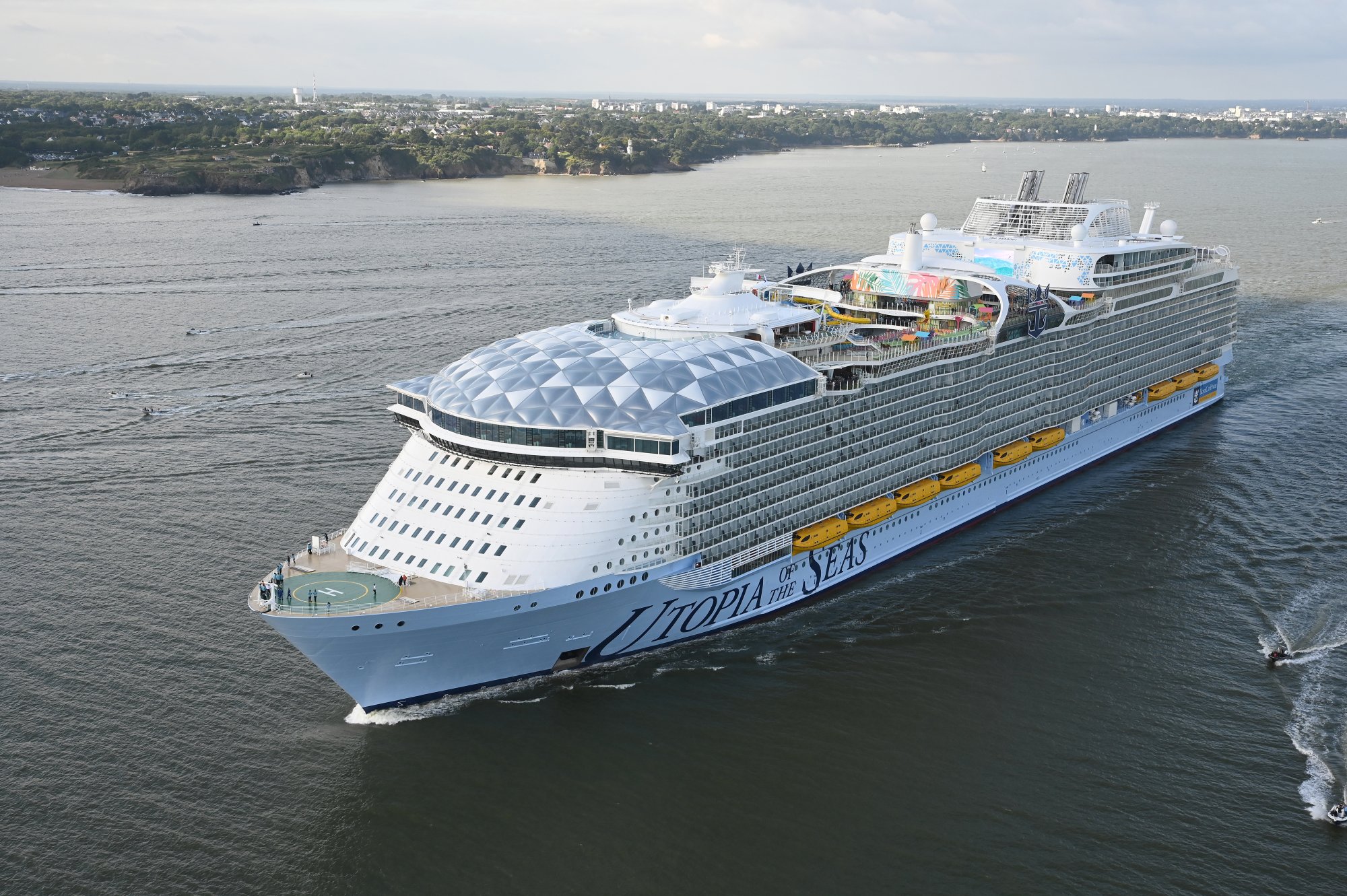A large modern cruise ship with distinctive light blue hull and white superstructure anchored in coastal waters, featuring a prominent geodesic dome structure on the forward deck and colorful entertainment areas visible amidships.