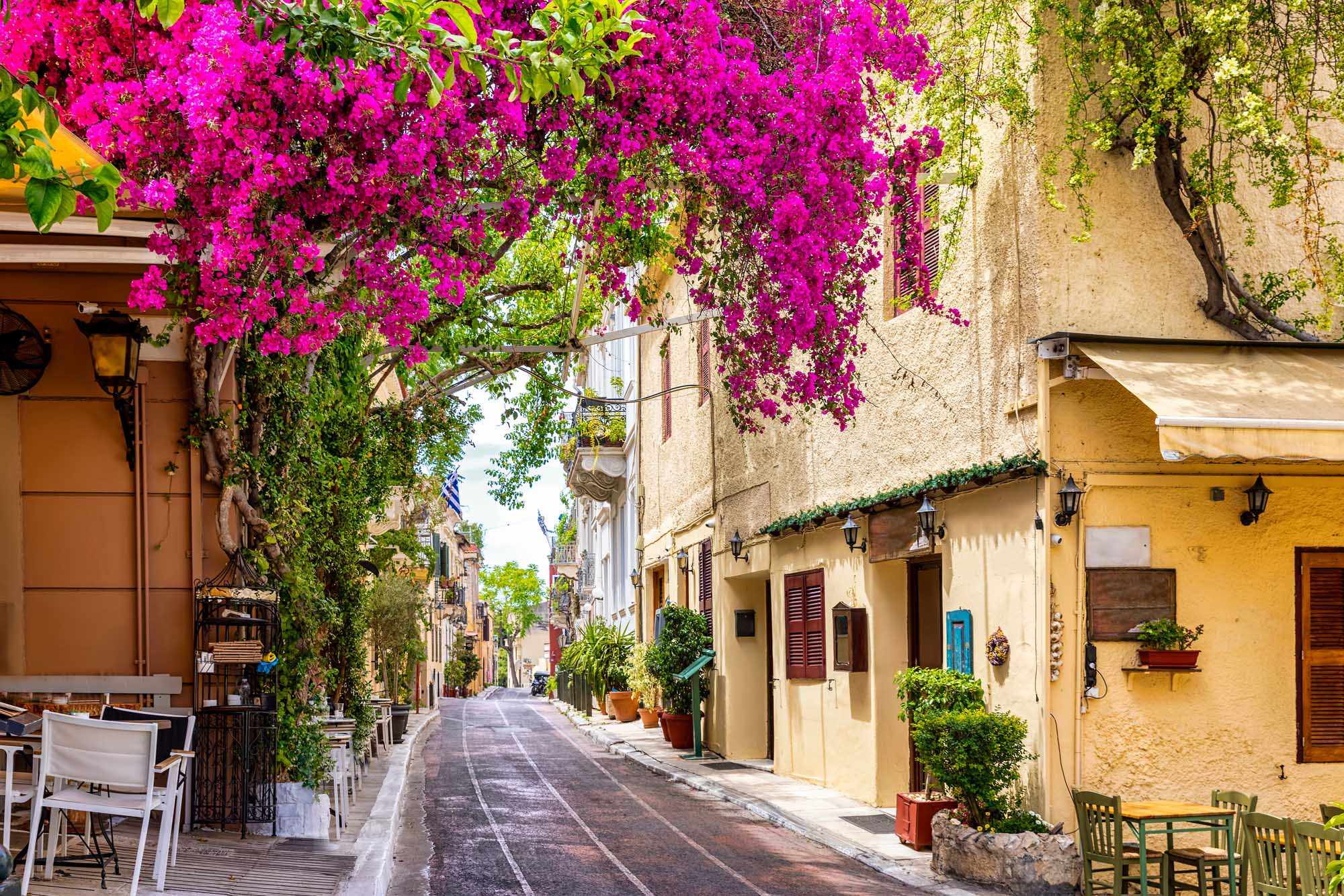 Vibrant Mediterranean Street with Bougainvillea A charming Mediterranean alleyway lined with traditional stone buildings and cascading magenta bougainvillea flowers overhead, featuring characteristic architecture with colorful doors, shutters, and outdoor cafe seating along a quiet street.