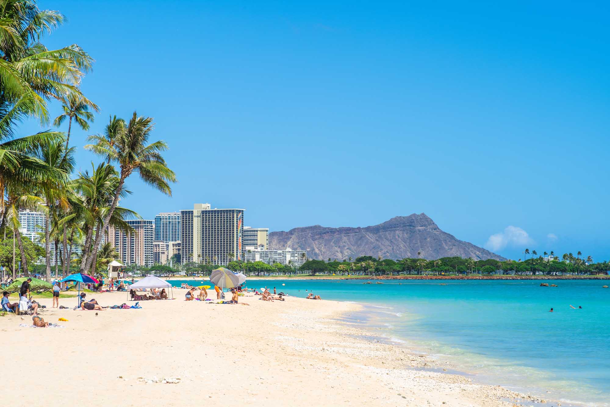 Waikiki Beach with Diamond Head Mountain A scenic view of Waikiki Beach in Honolulu, Hawaii, featuring pristine white sand, crystal-clear turquoise waters, and beachgoers enjoying the tropical weather. The iconic Diamond Head mountain rises majestically in the background.