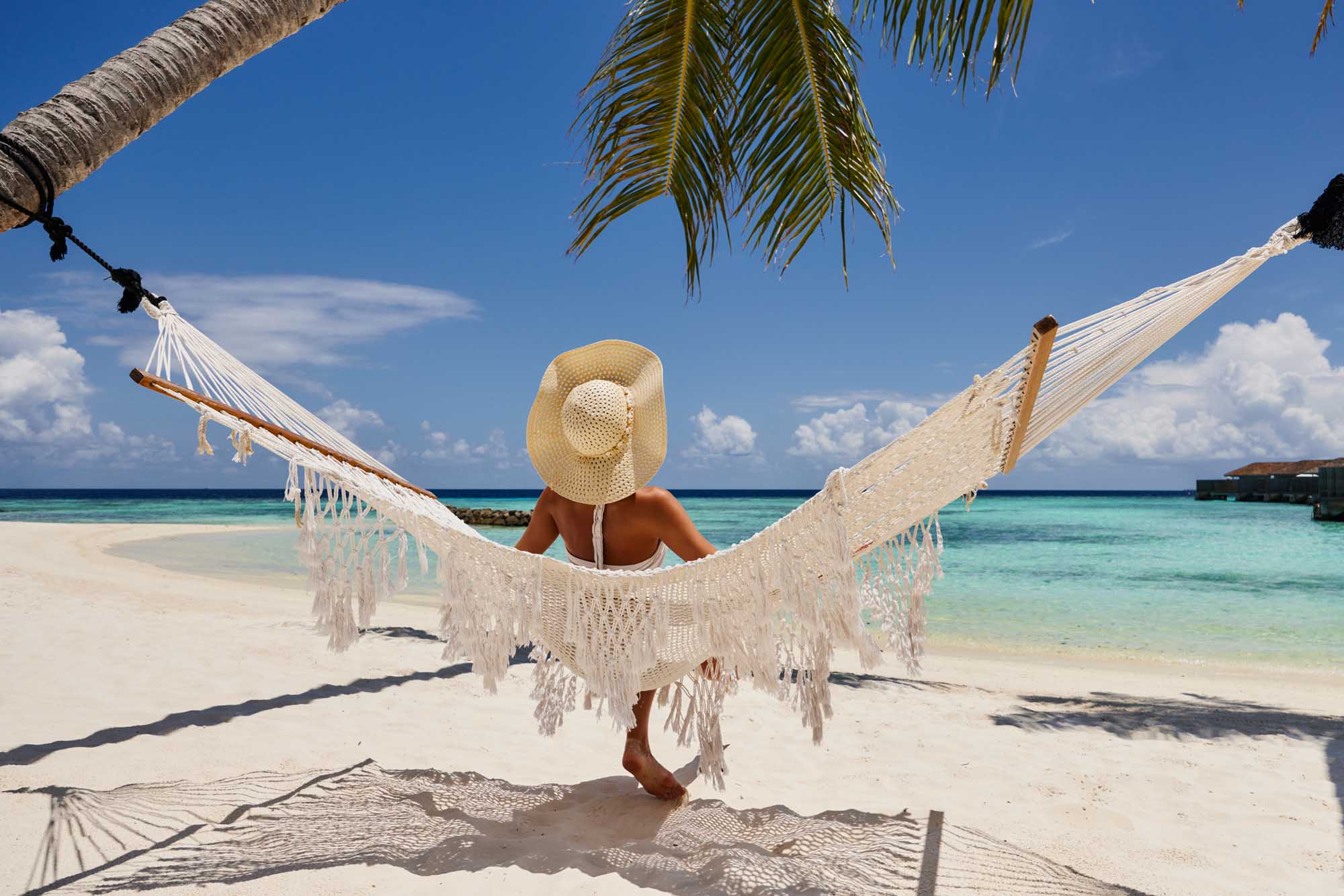 Woman Relaxing in Hammock on Tropical Beach A rear view of a woman wearing a straw sun hat relaxing in a white macrame hammock strung between palm trees on a pristine tropical beach, overlooking turquoise waters and a clear blue sky dotted with white clouds.