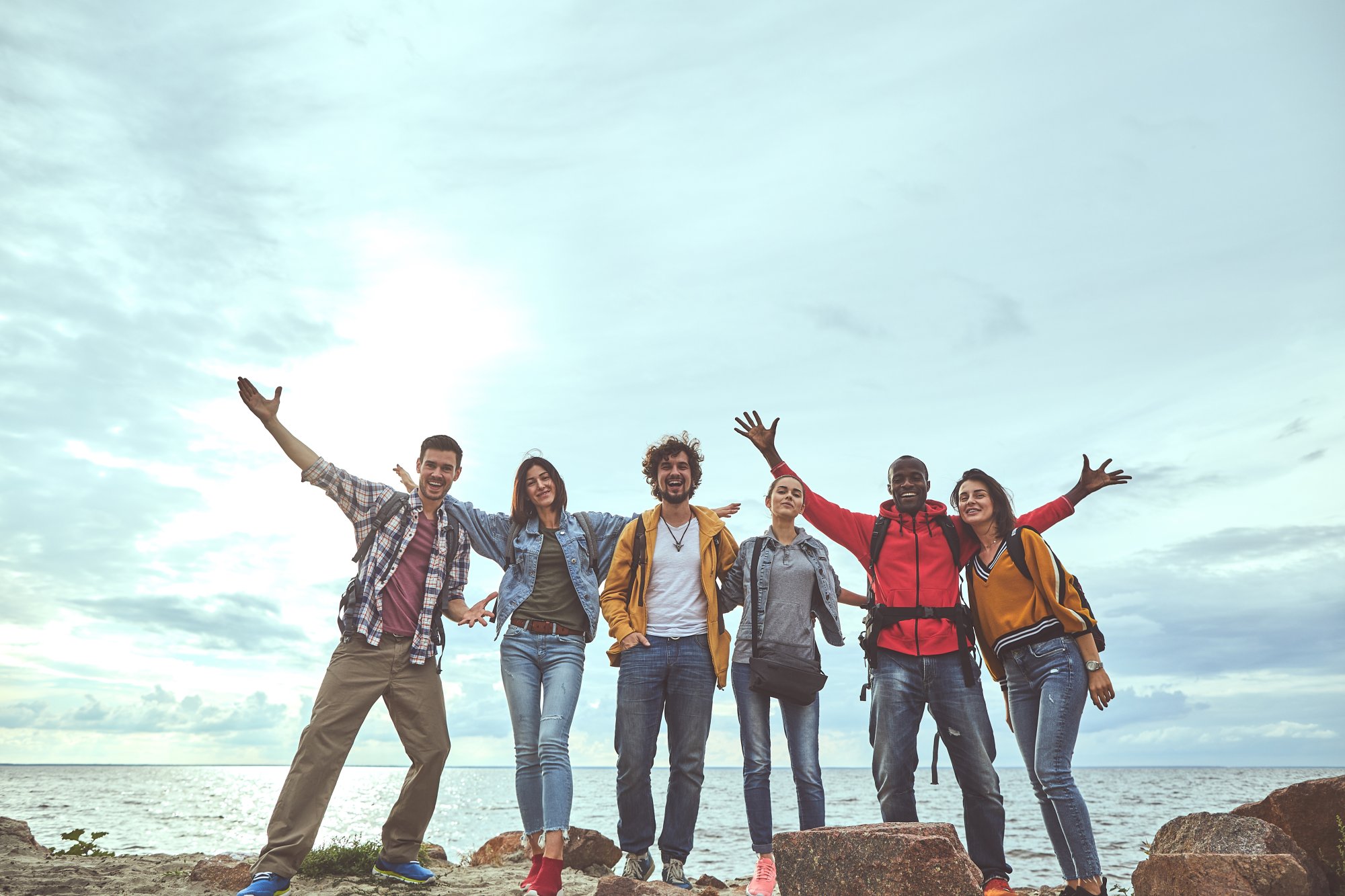 A diverse group of six young adults standing on rocks by the ocean with their arms raised in celebration and joy during a travel adventure outing.